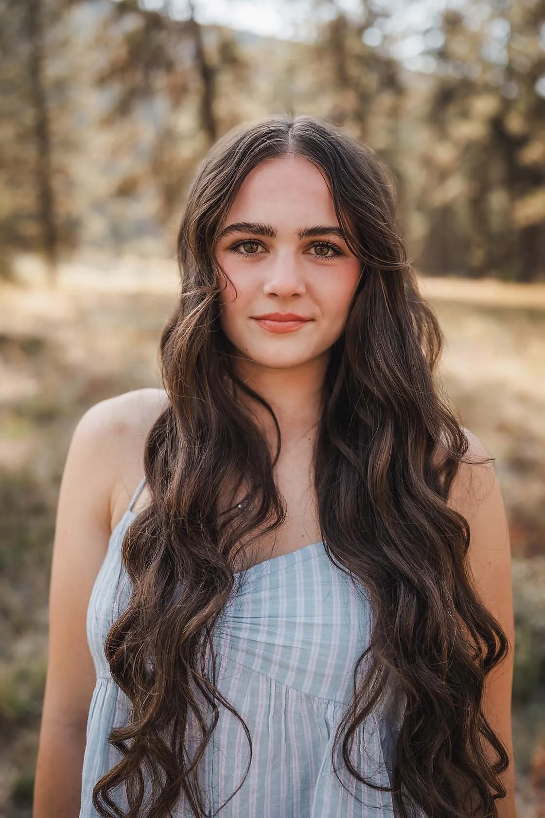 High school senior girl with long wavy hair in a light blue dress standing in a sunlit forest, garden valley Idaho senior photographer