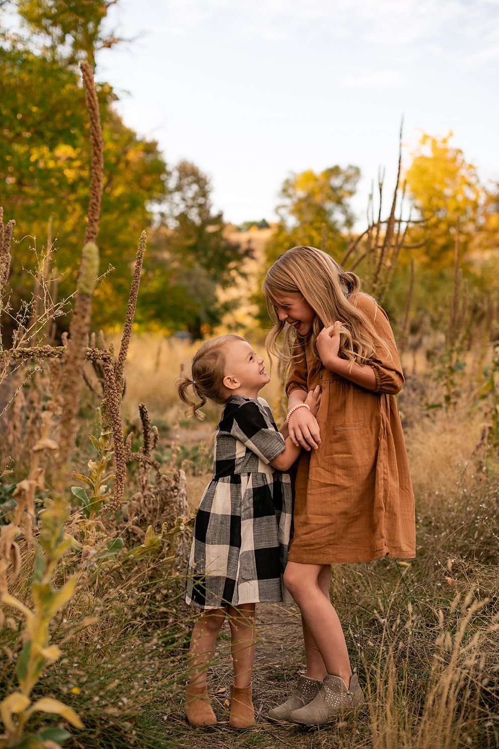Two sisters smiling at each other in a Boise meadow during a fall family photo session, wearing plaid and rust-colored dresses