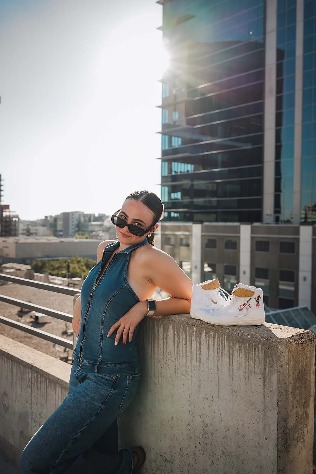 Eagle High School senior wrestler in denim and sunglasses leaning on a concrete ledge next to her wrestling shoes with downtown Boise Idaho buildings behind her