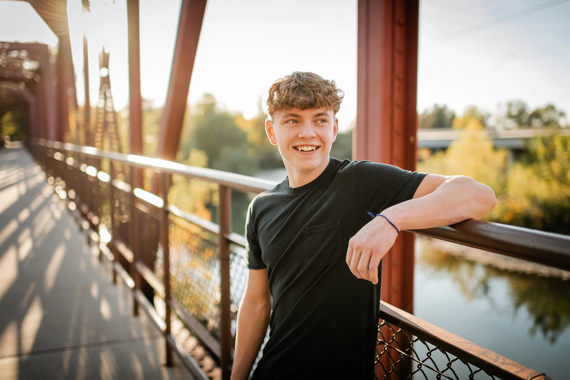 Senior boy in black t-shirt leaning on red bridge railing at golden hour in Boise, Idaho