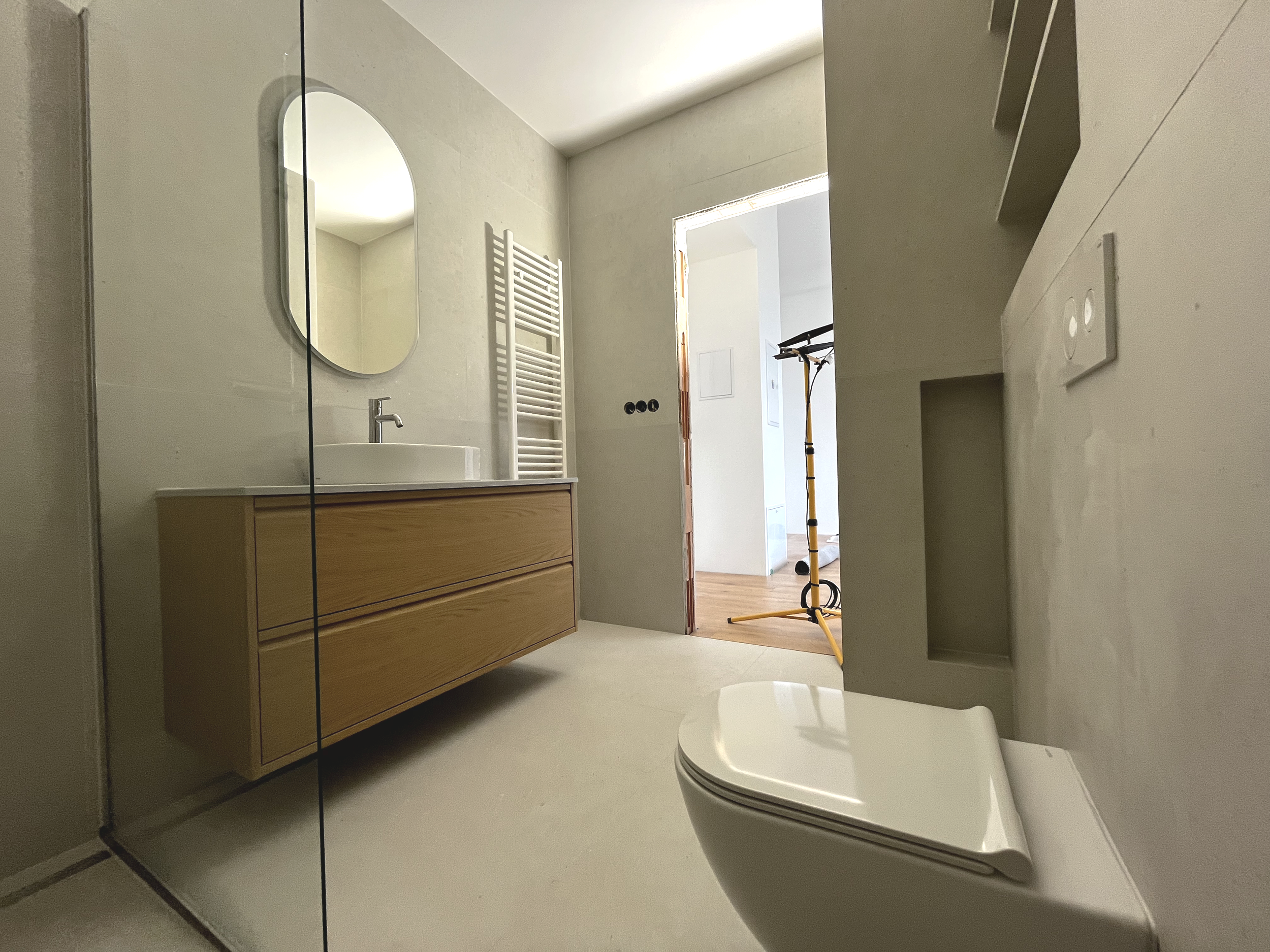 Modern bathroom with light-colored walls, wooden vanity with a white vessel sink, oval mirror, white heated towel rack, and a white toilet, with a doorway leading to a room with a black and yellow floor lamp.