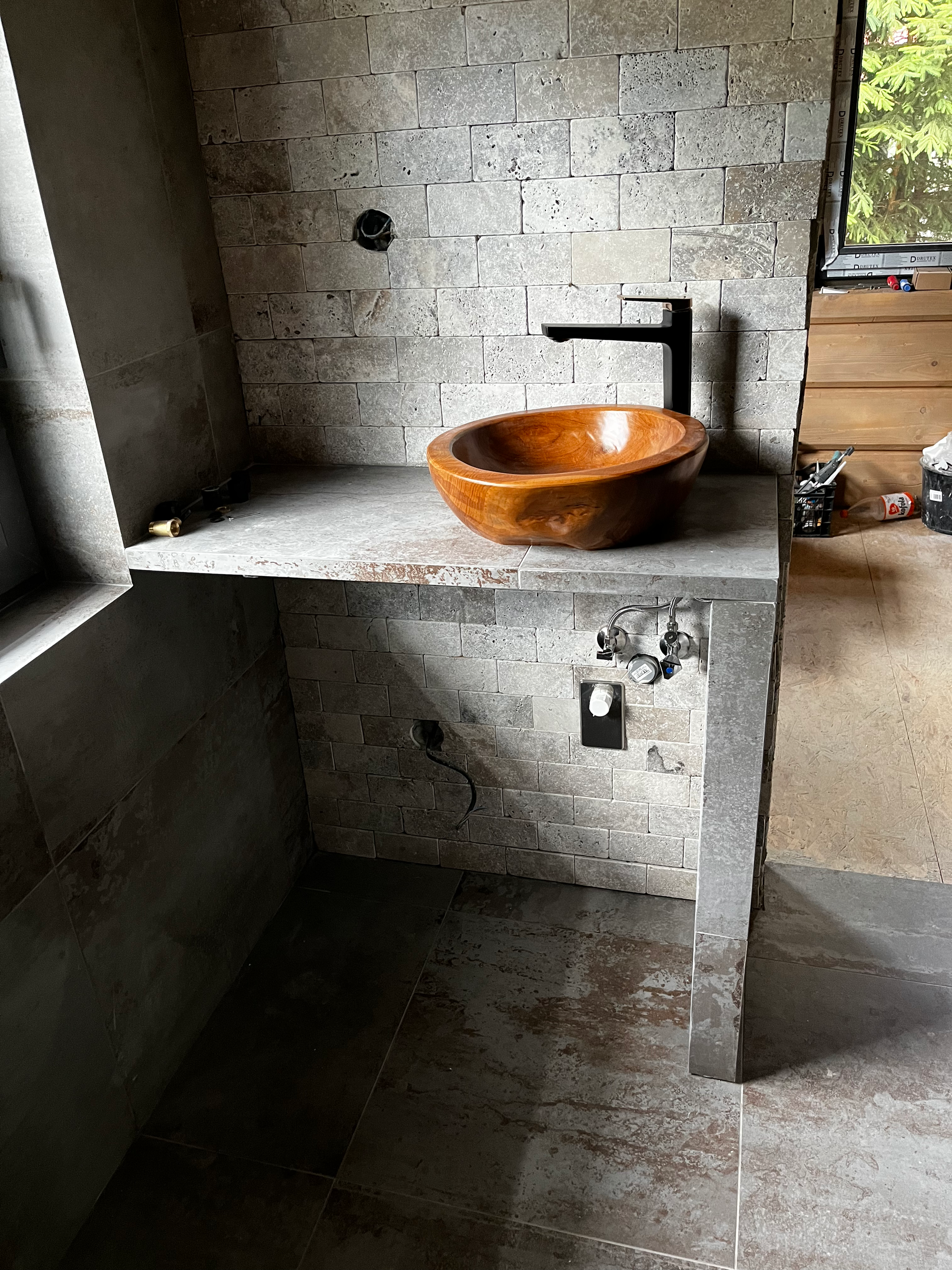 An unfinished bathroom vanity with a round wooden bowl sink, black faucet, and gray brick tiles on the wall and countertop. Plumbing connections are visible below, and construction tools and supplies are scattered around.