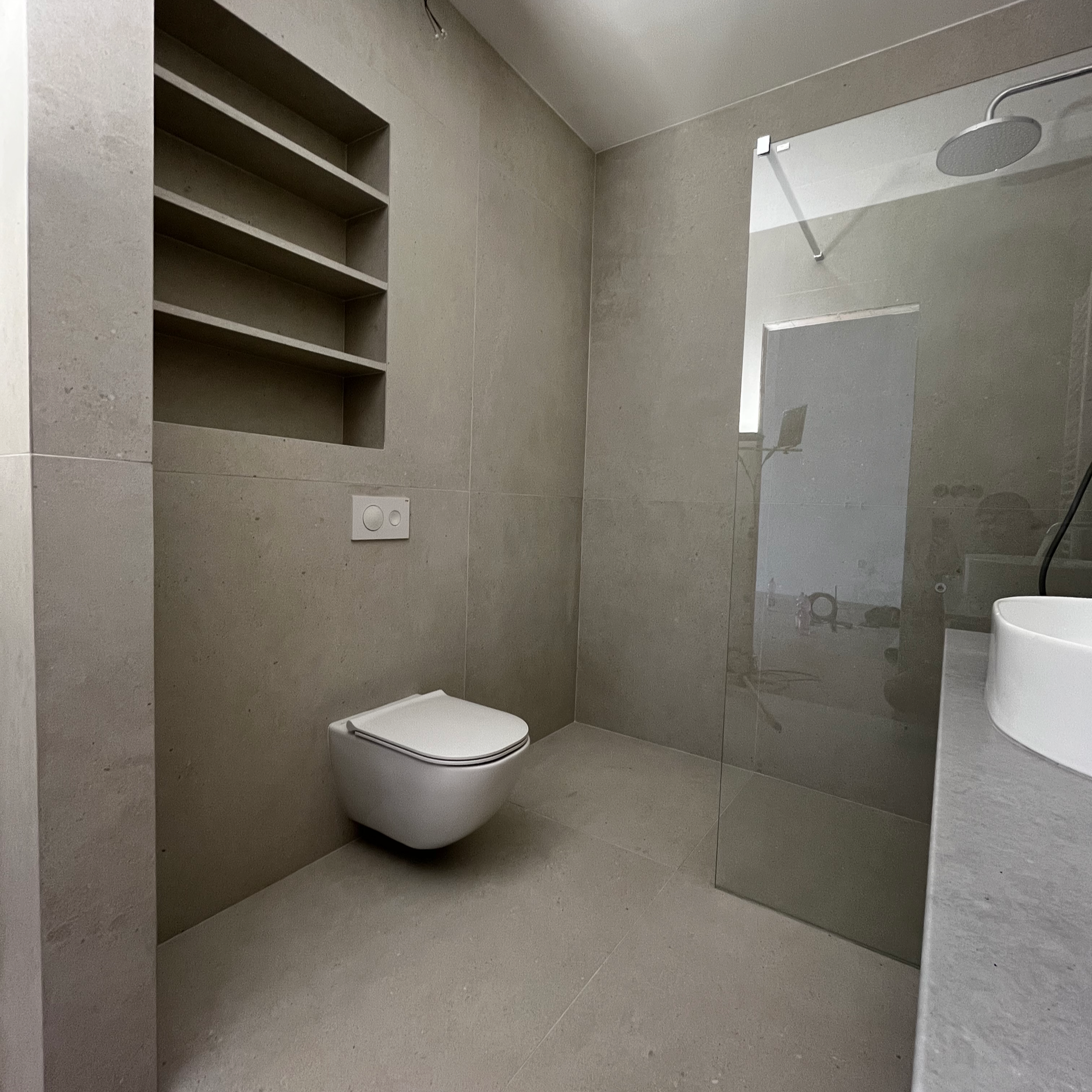 Modern bathroom with a wall-mounted toilet, built-in wall shelves, a large shower area with a glass partition, and a countertop with a vessel sink.