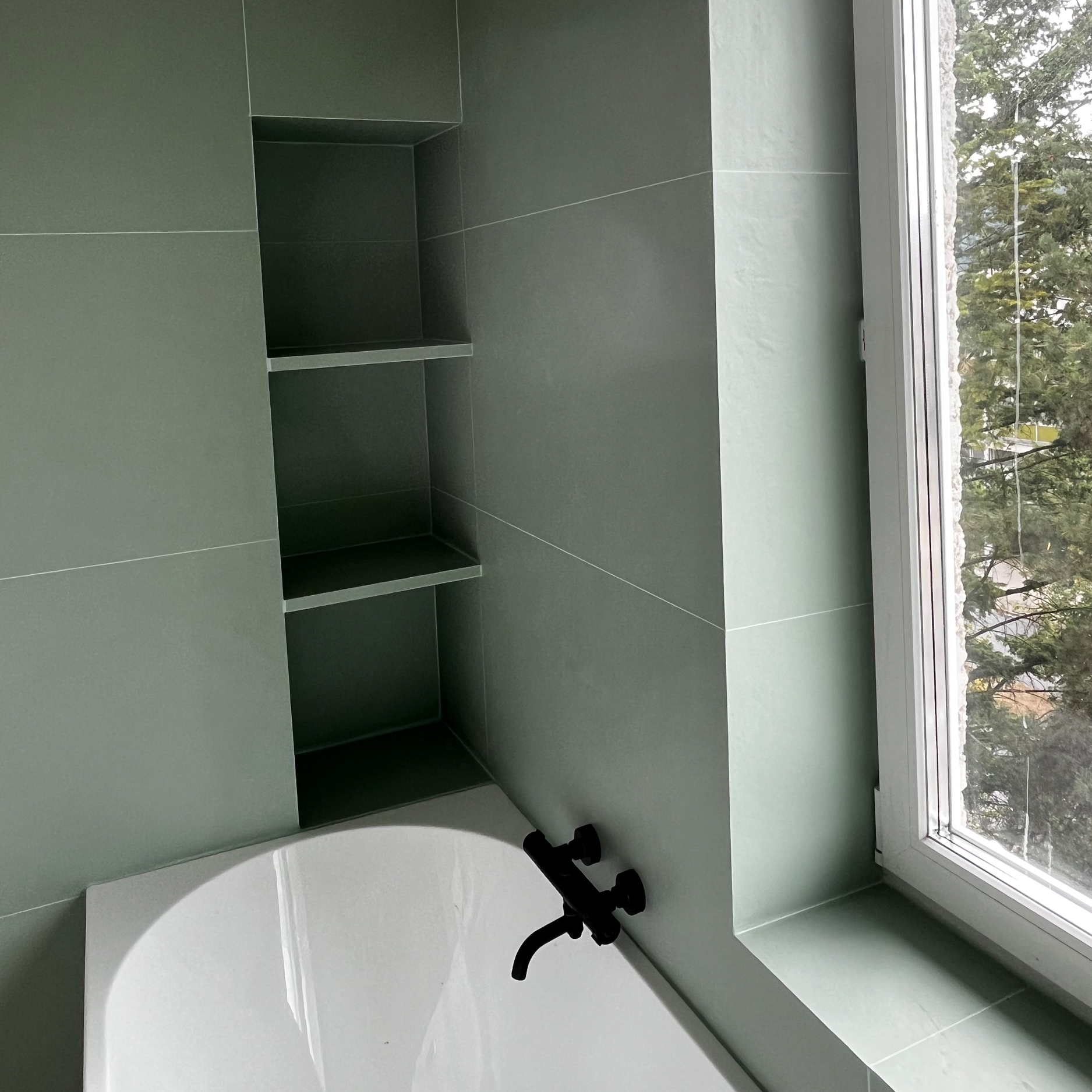Green-tiled bathroom with a window showing trees outside, black faucet, and built-in shelving