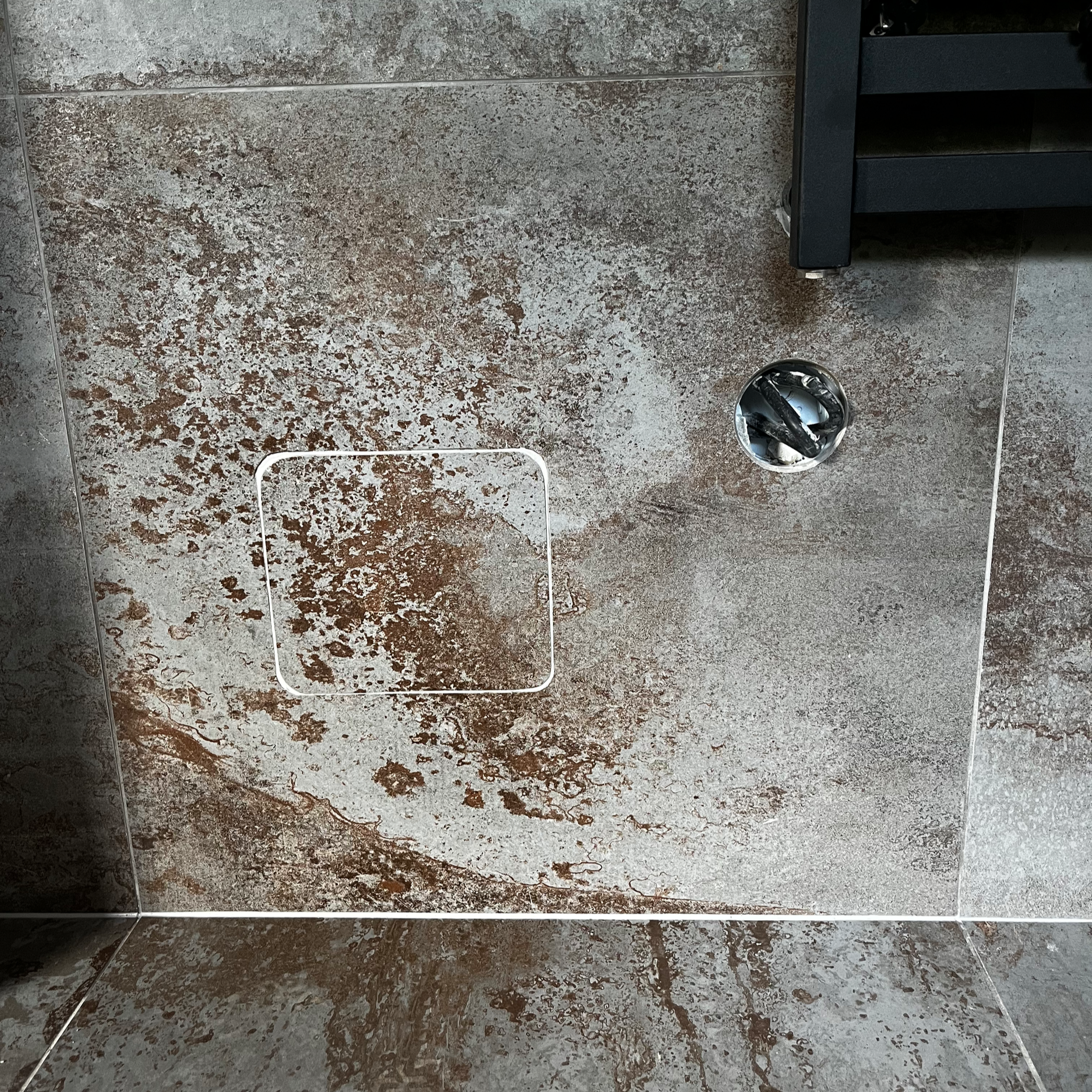 View of a tiled bathroom wall and floor with a metal drain cover and a black shelf. The tiles have a brown and gray marbled pattern.