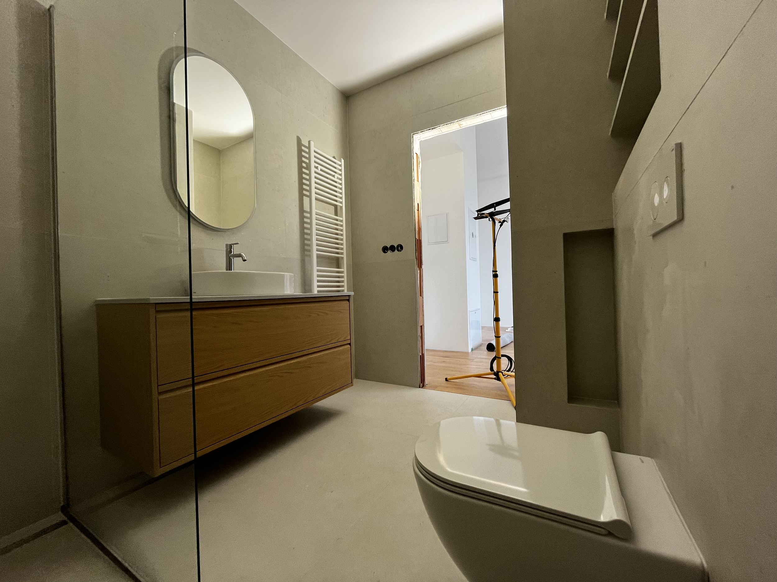 A modern bathroom with a wooden vanity, an oval mirror, a white sink, a heated towel rail, and a toilet. There is a doorway leading to an adjoining room with a tripod or light stand and a glimpse of wooden flooring.