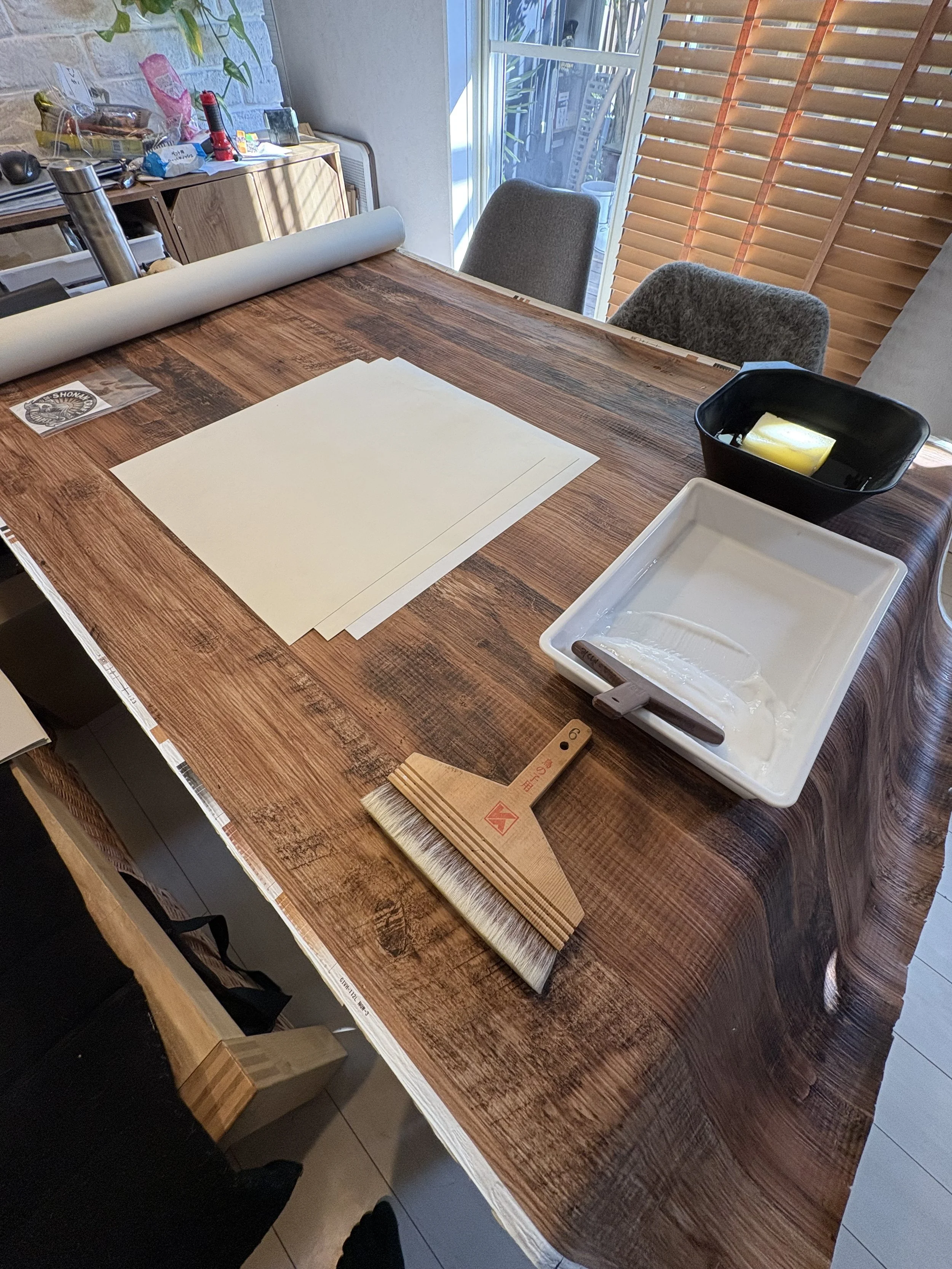 テーブルの上に壁紙と刷毛、白いトレイが置かれた作業スペース。背景に窓と日差し。