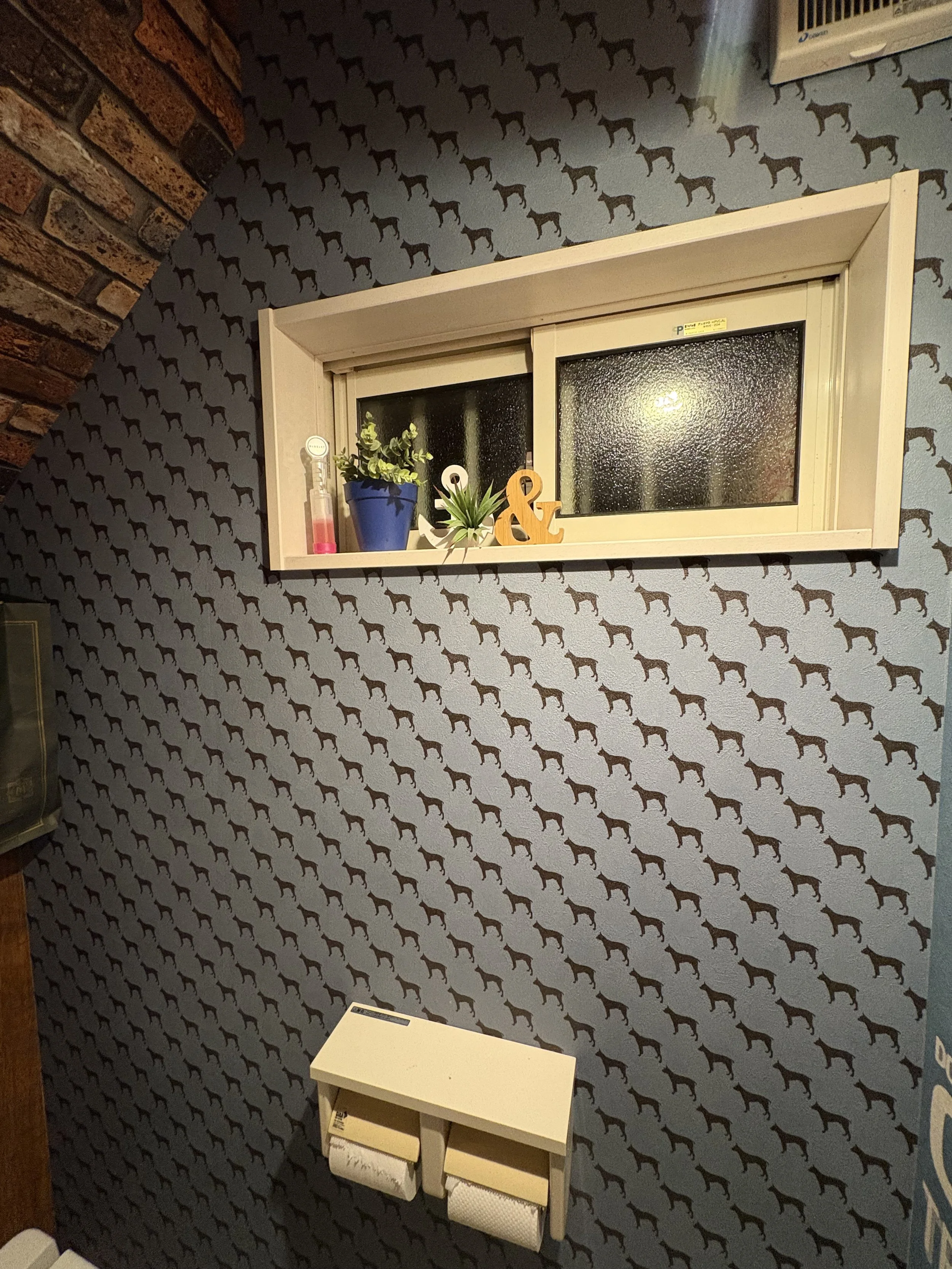 トイレの壁には犬の模様の壁紙があり、小窓には植物と装飾物が置かれている。壁にはトイレットペーパーホルダーが設置されている。