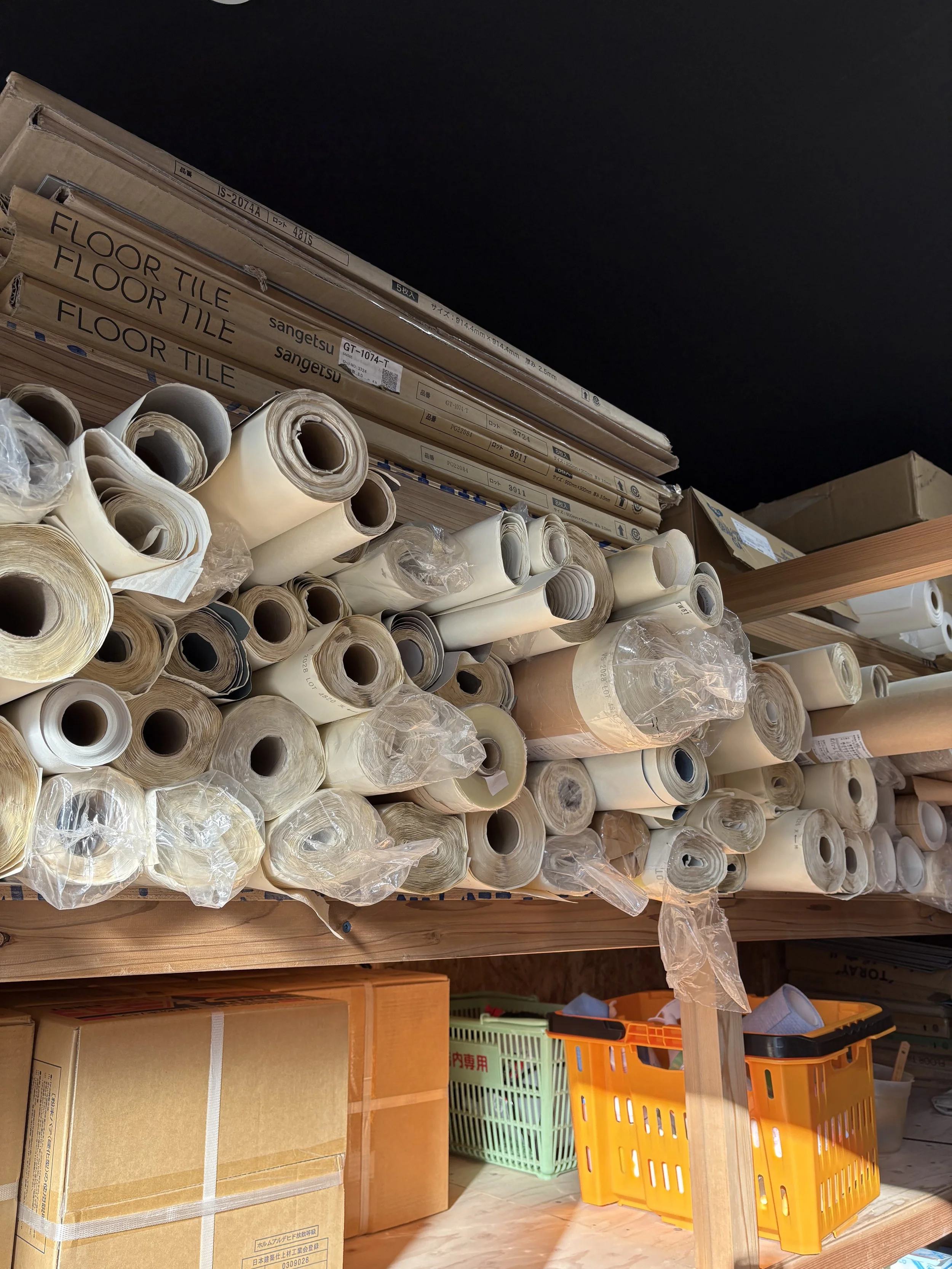 棚には、フローリングタイルの箱やロール状のビニール素材が収納されている。下には段ボール箱とプラスチックの収納バスケットが置かれている。