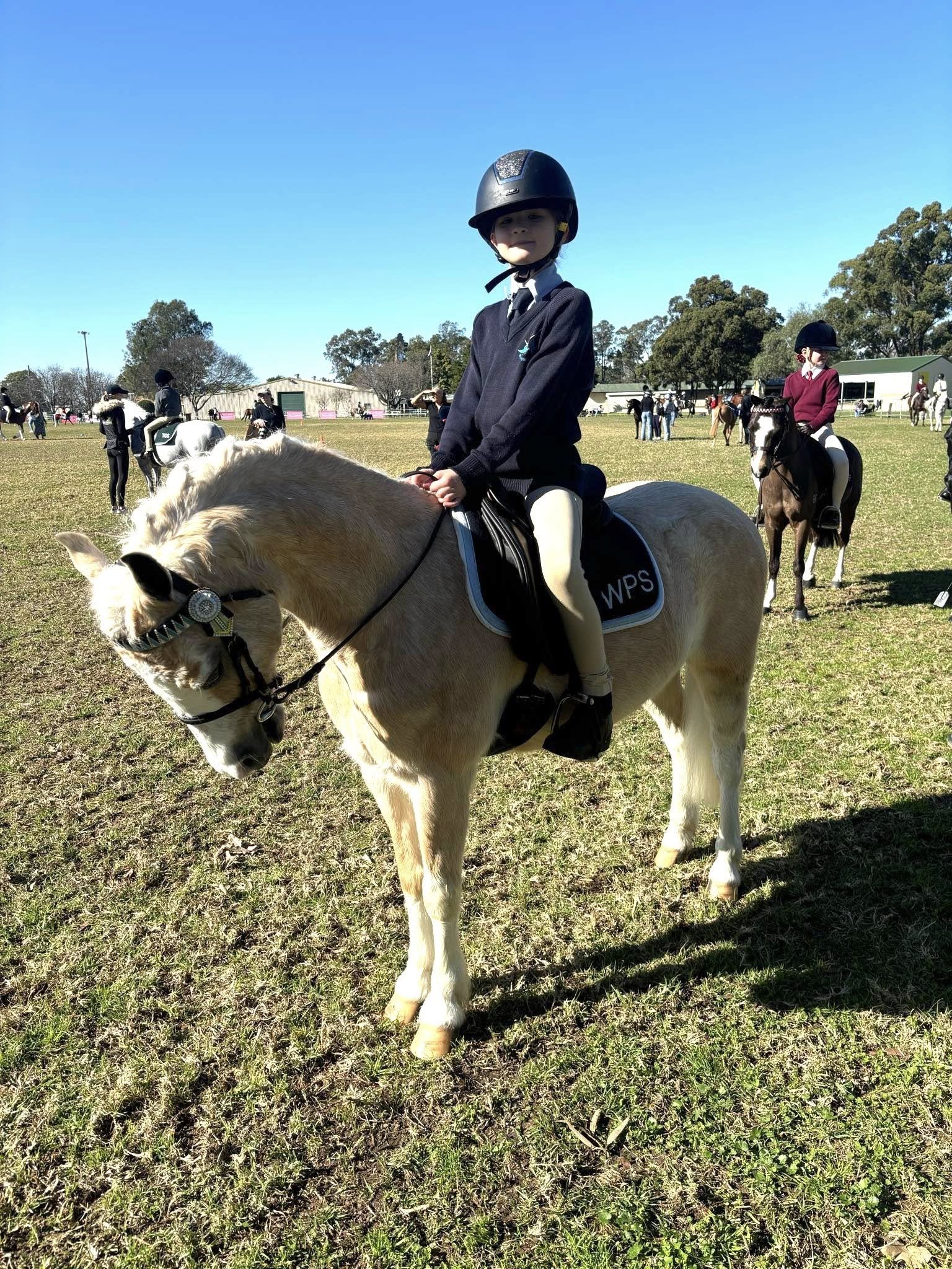 A young girl wearing a helmet riding a white horse on a grassy field during a riding event. Other riders and spectators are visible in the background on a bright, sunny day.