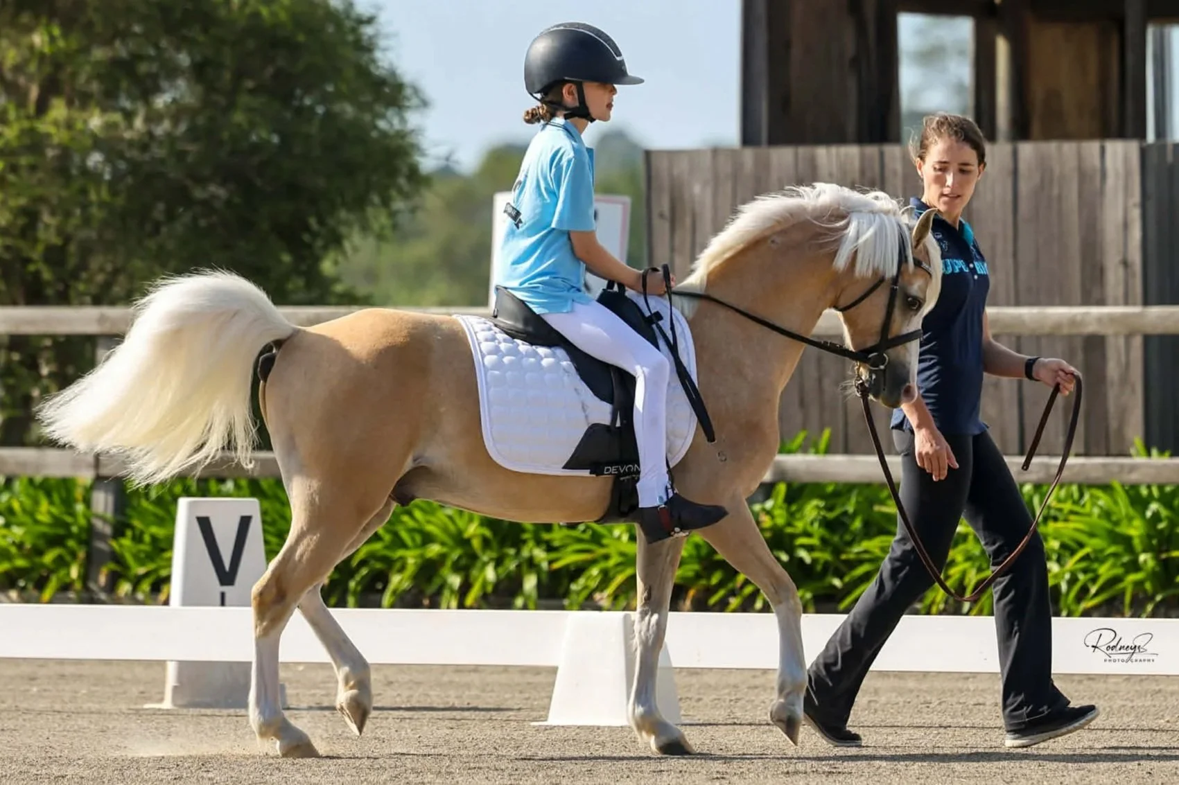 A young girl riding a light-colored pony in an outdoor riding arena, guided by a woman. The girl wears a helmet and a blue shirt.