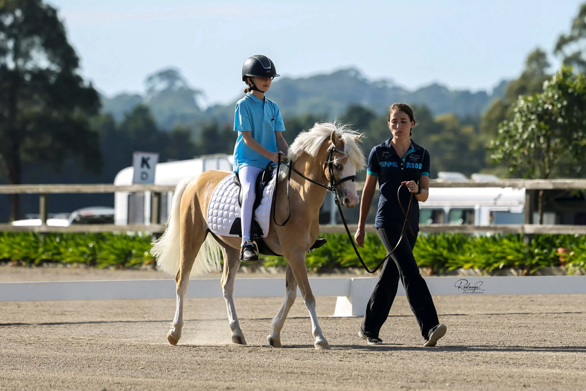 A young girl riding a light-colored pony with a white mane at an outdoor equestrian facility, guided by a woman in dark clothing.