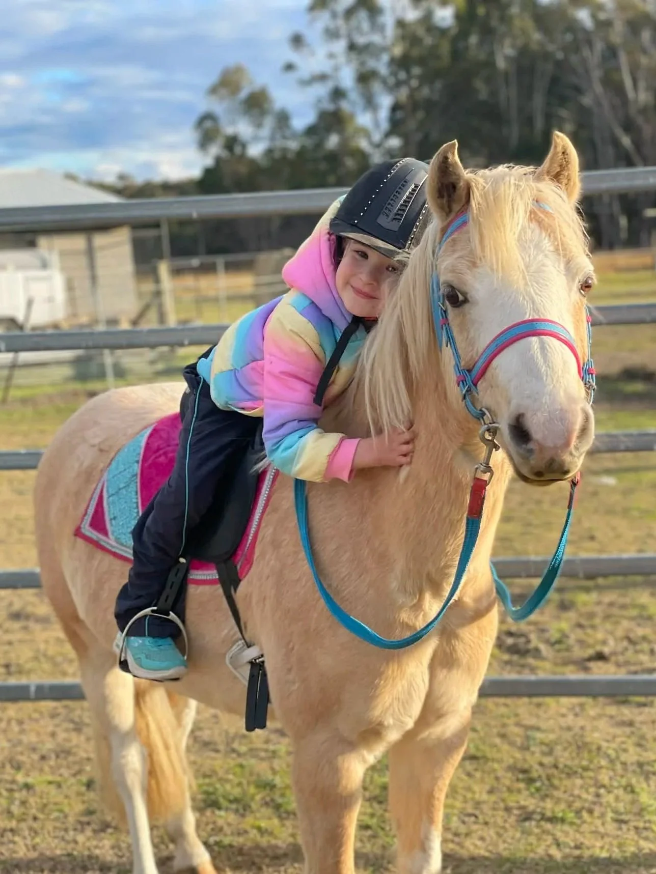 A young girl with a colorful hoodie smiling while hugging a cream-colored horse with a pink and blue halter, standing in a fenced outdoor area on a partly cloudy day.