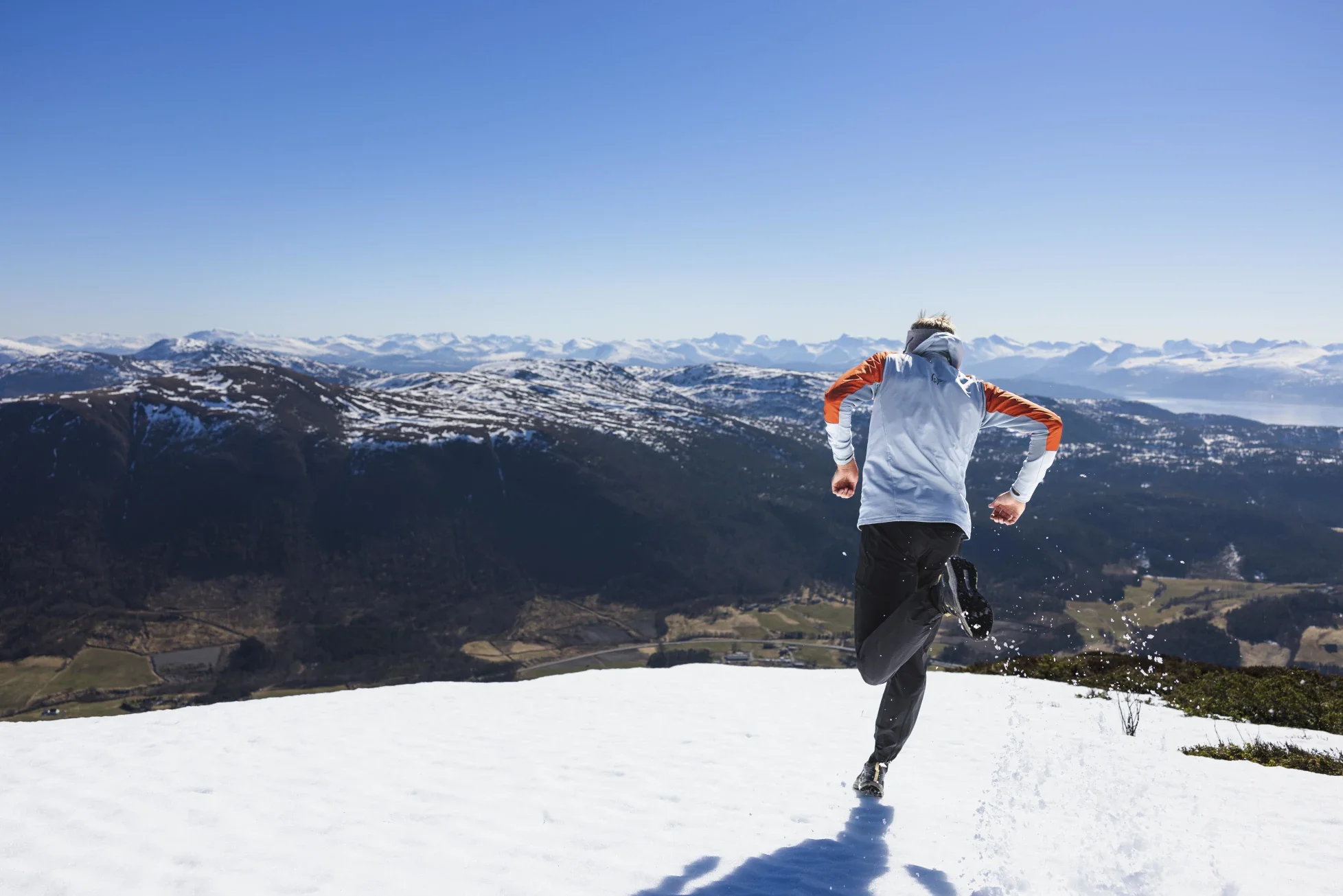 A person running on snow-covered ground with mountains and blue sky in the background.