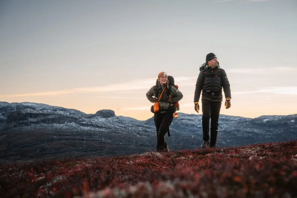 Two women hiking in a mountainous landscape at sunset, wearing outdoor gear and backpacks.