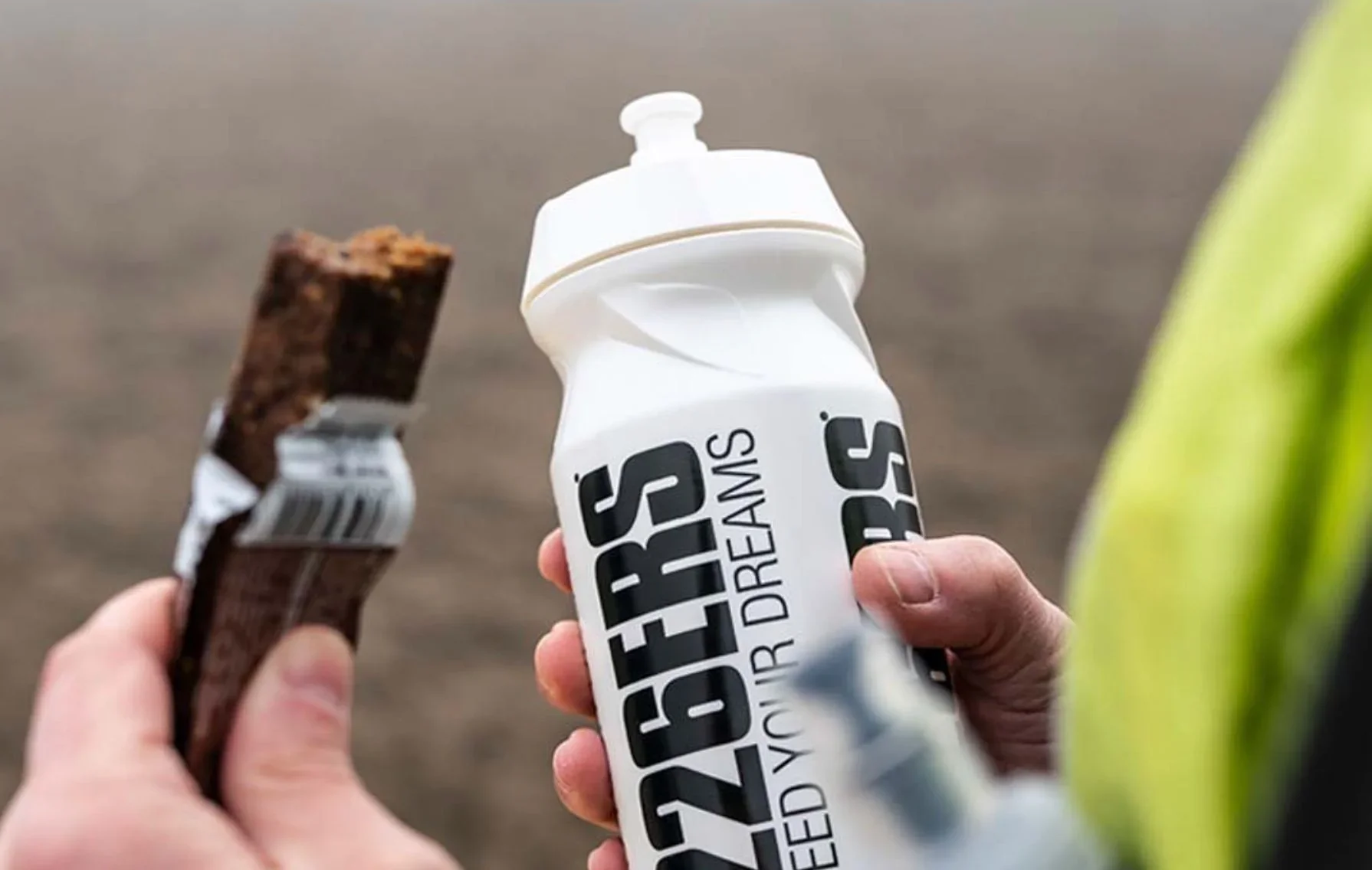A person holding a chocolate protein bar in one hand and a bottle of Gore® Vapor Dry Stream in the other, with a blurred outdoor background.