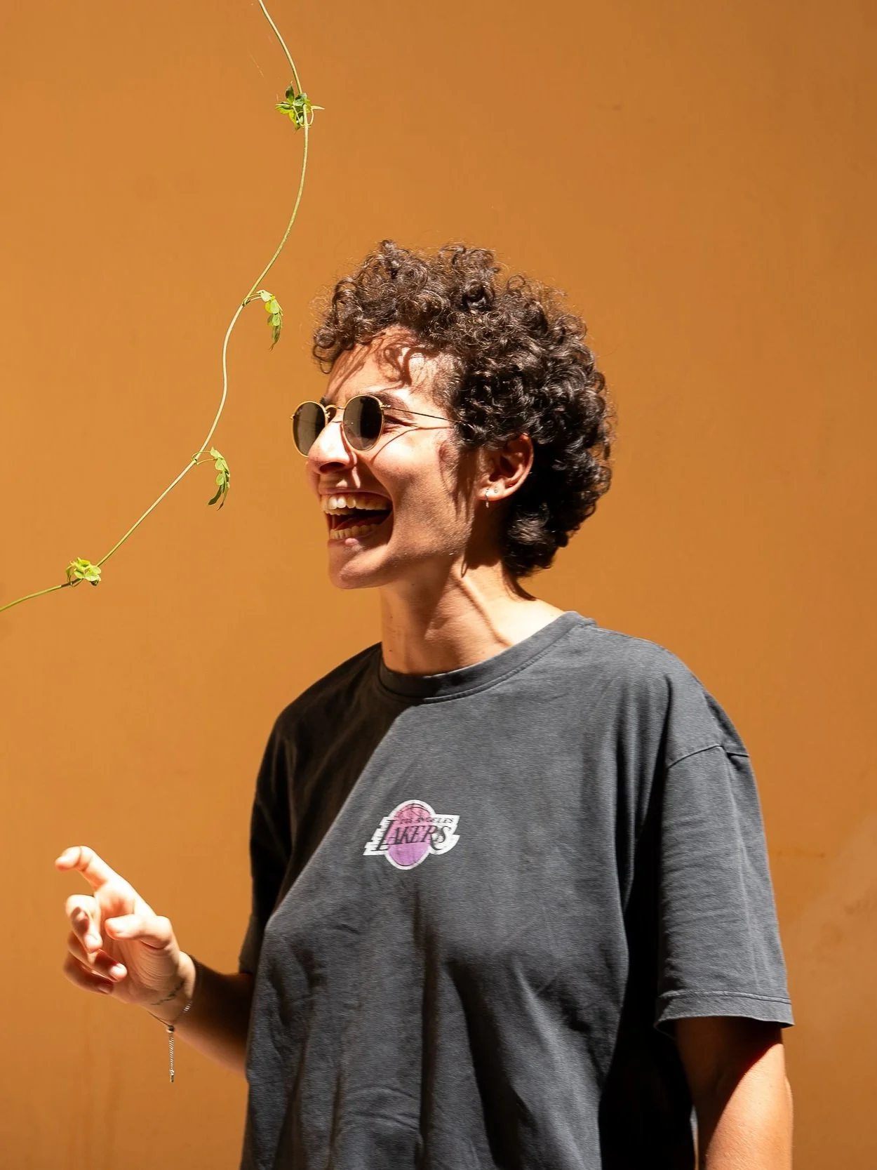 A woman with short curly hair wearing sunglasses and a black T-shirt with a Lakers logo, smiling and pointing, against a plain tan wall.