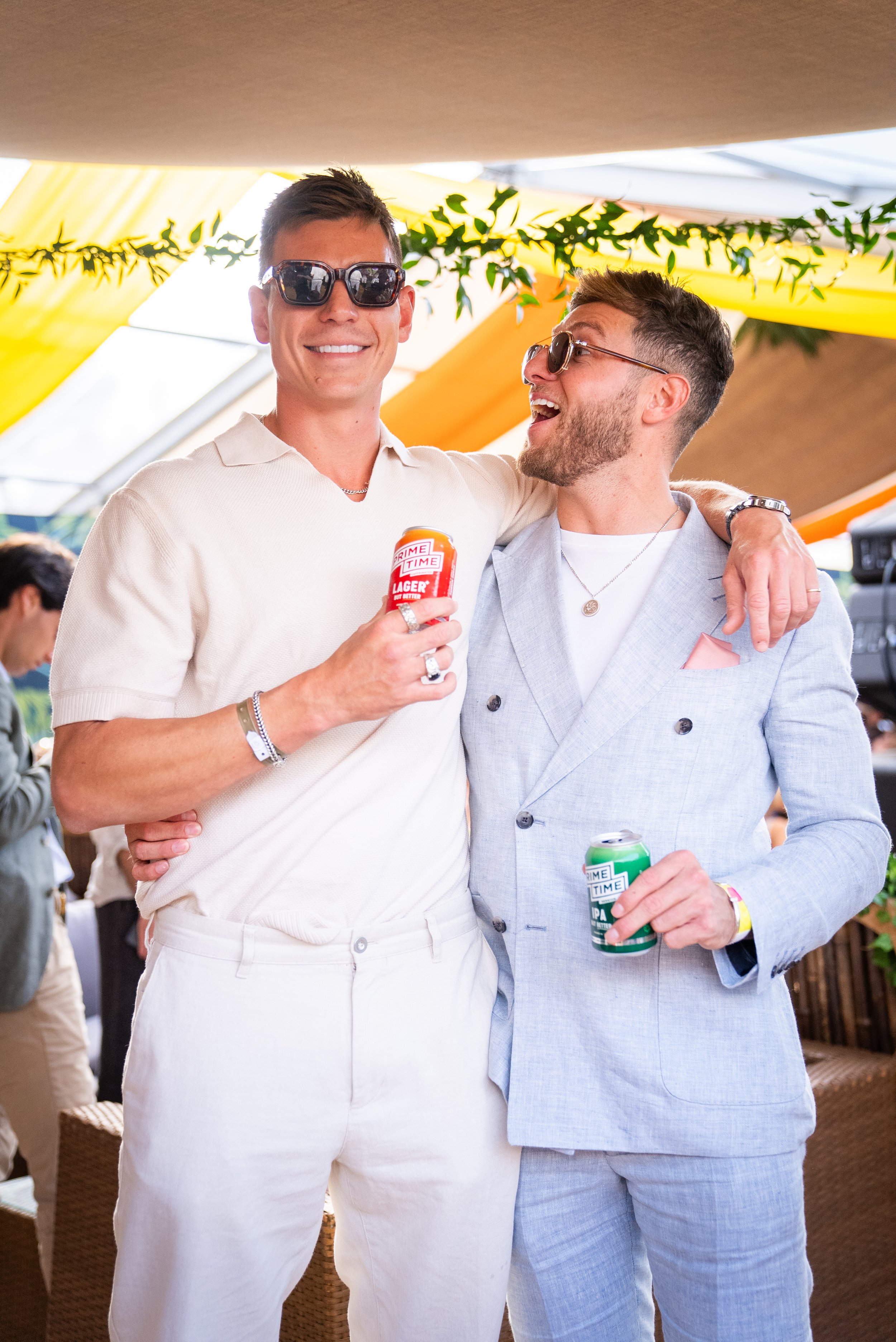 Tristan Phipps and friend in stylish outfits at an event, smiling and holding canned drinks, with one wearing a white collared shirt and the other in a light blue blazer, standing under a canopy with decorative greenery.