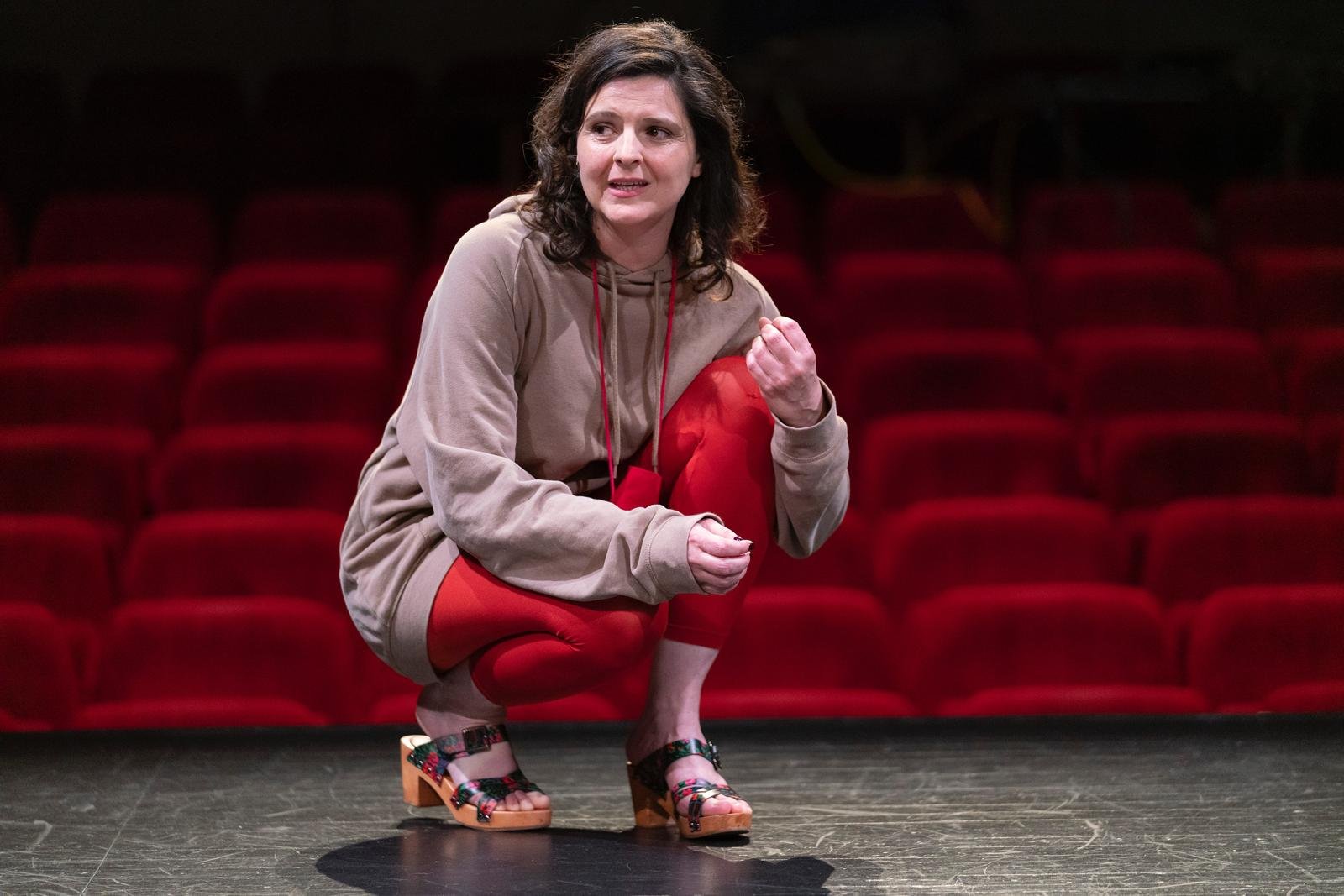 Une femme accroupie sur une scène de théâtre avec un fond de sièges rouges.