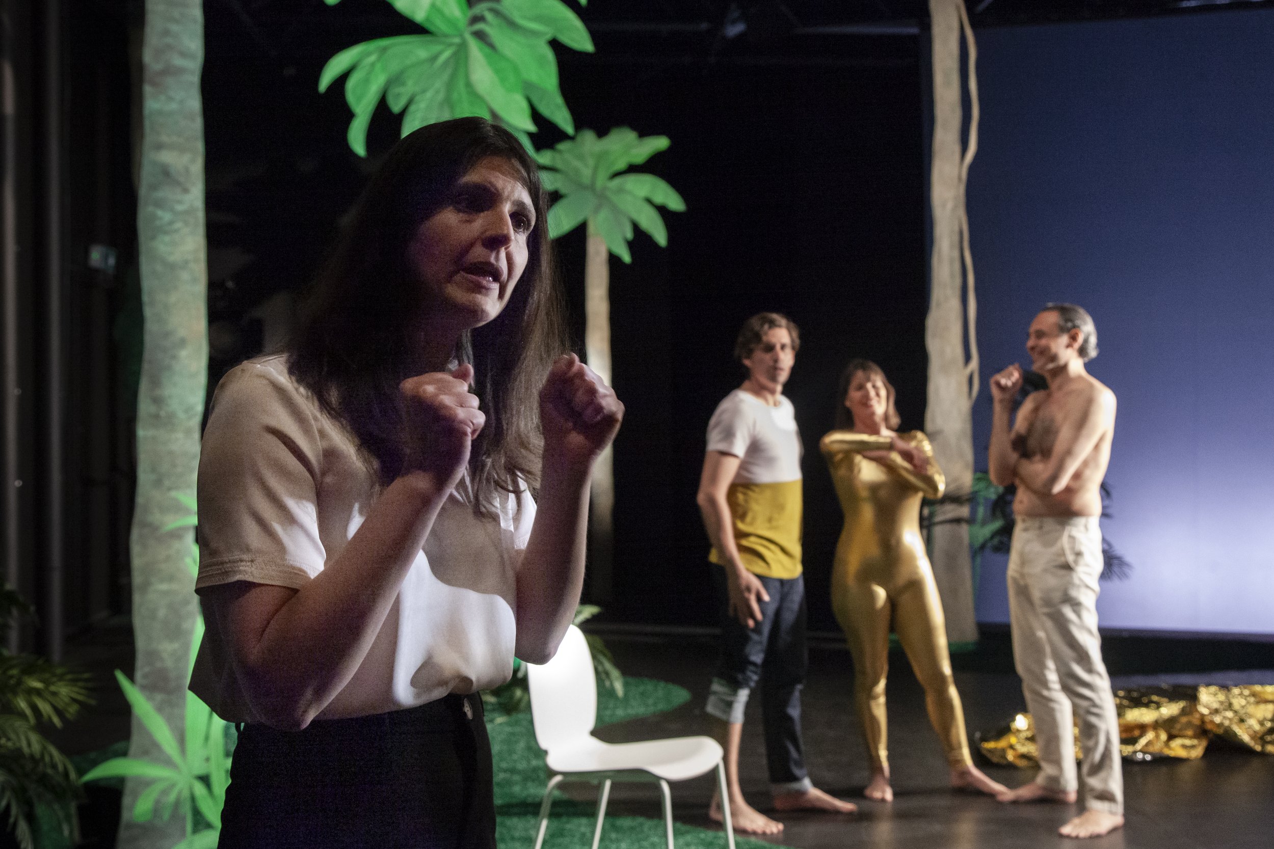Scène de théâtre avec quatre acteurs, dont une femme au premier plan qui semble exprimer une émotion intense. Trois autres acteurs en arrière-plan, dont un homme torse nu et une femme en combinaison dorée, dans un décor de jungle avec des arbres arti
