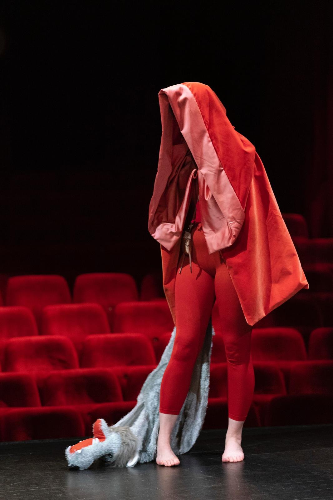 Personne en costume rouge avec capuche, traînant une peluche ressemblant à un loup, sur une scène devant des sièges rouges.