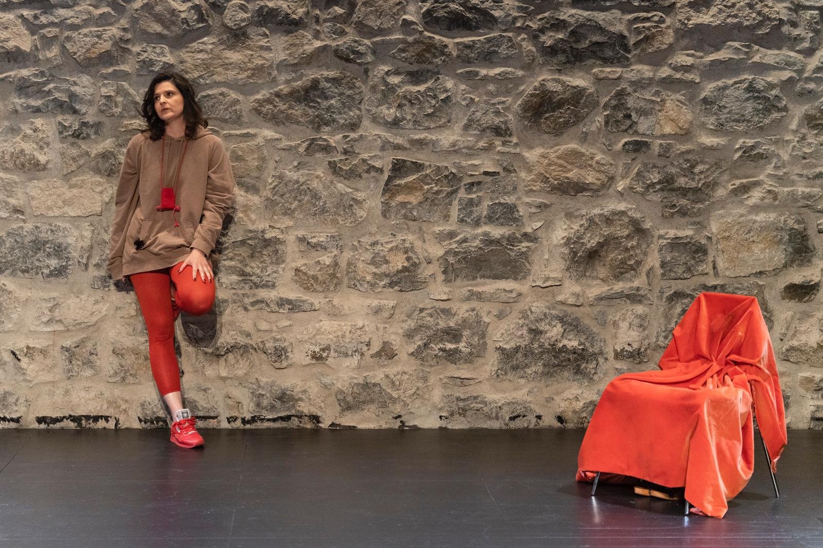 Une femme appuyée contre un mur en pierre avec une chaise recouverte d'un drap rouge à côté d'elle.