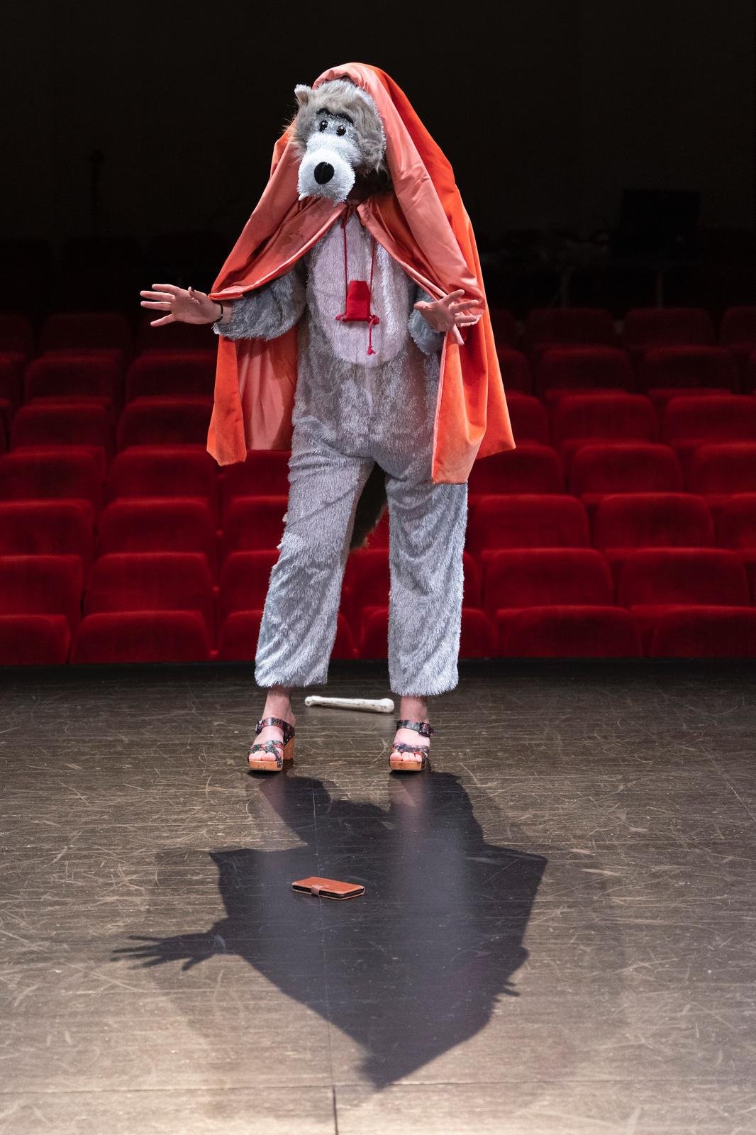 Une personne costumée en loup avec une cape rouge sur une scène de théâtre, devant un public assis sur des sièges rouges.