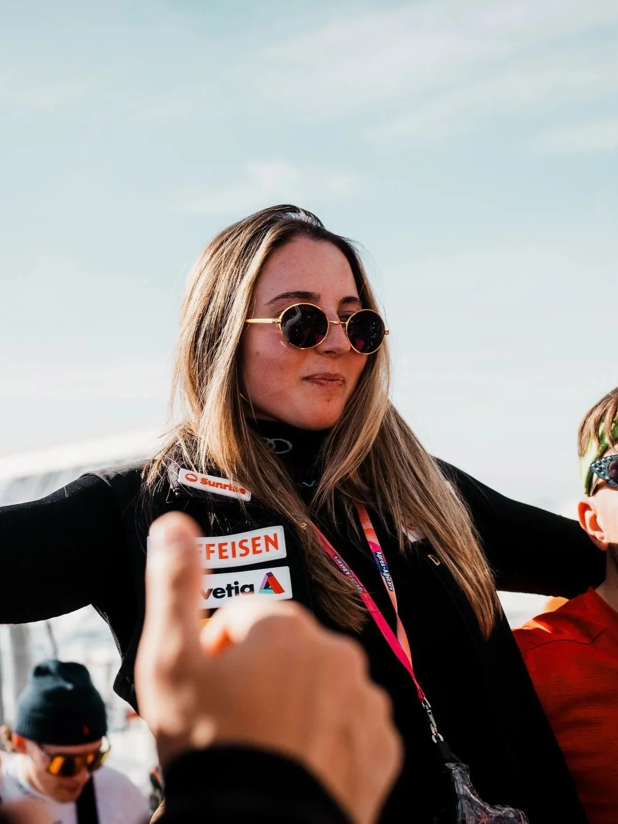 Isabella mit Sonnenbrille und langen Haaren trägt eine schwarze Jacke mit Logos. Sie hebt die Arme in der Luft. Im Hintergrund sind verschwommen andere Personen zu sehen.