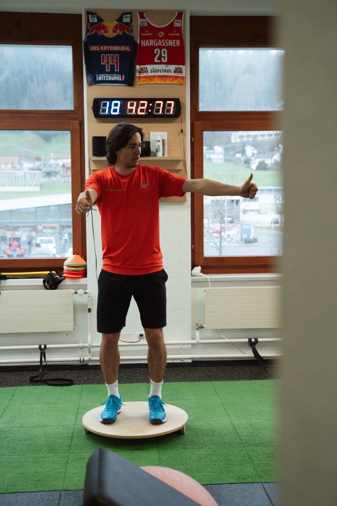 Mann in rotem Shirt macht Balanceübung auf einer runden Plattform in einem Fitnessstudio.