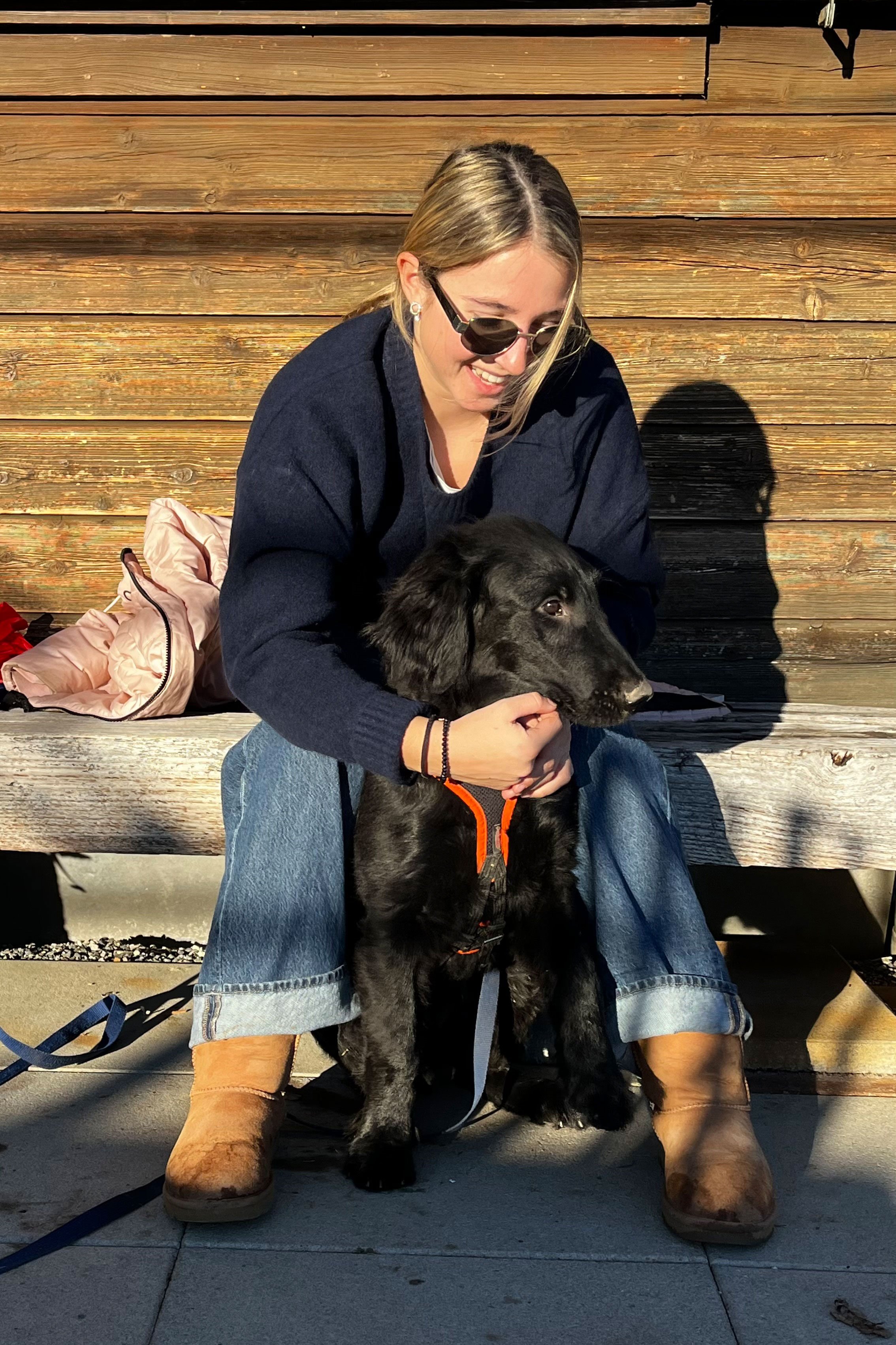 Isabella sitzt auf Bank mit Hund im Freien, Sonnenbrille, Jeans, Holzplanken im Hintergrund.