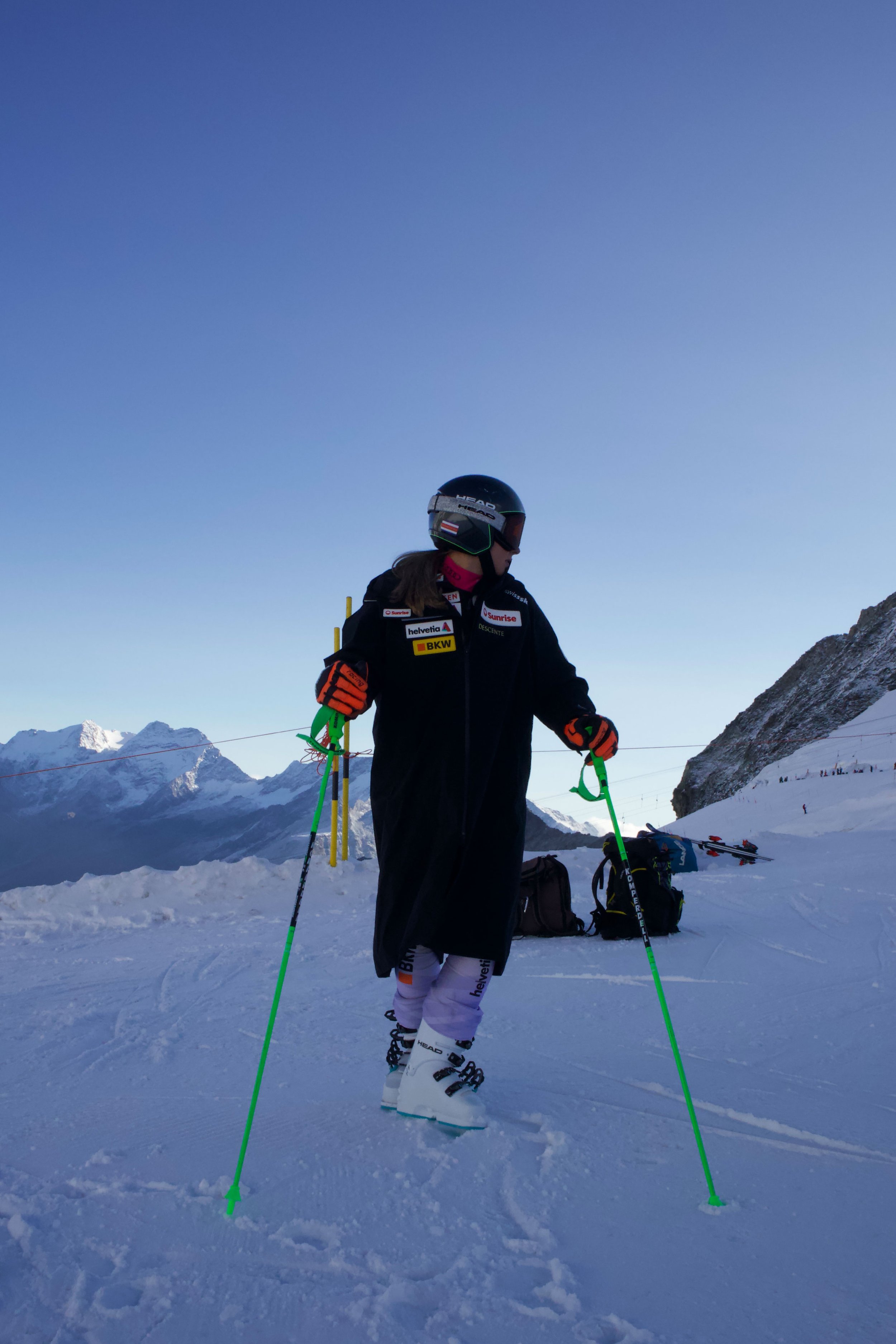 Skifahrer auf einem schneebedeckten Berg mit Skistöcken, Skischuhen und Helm, im Hintergrund Berge und blauer Himmel.
