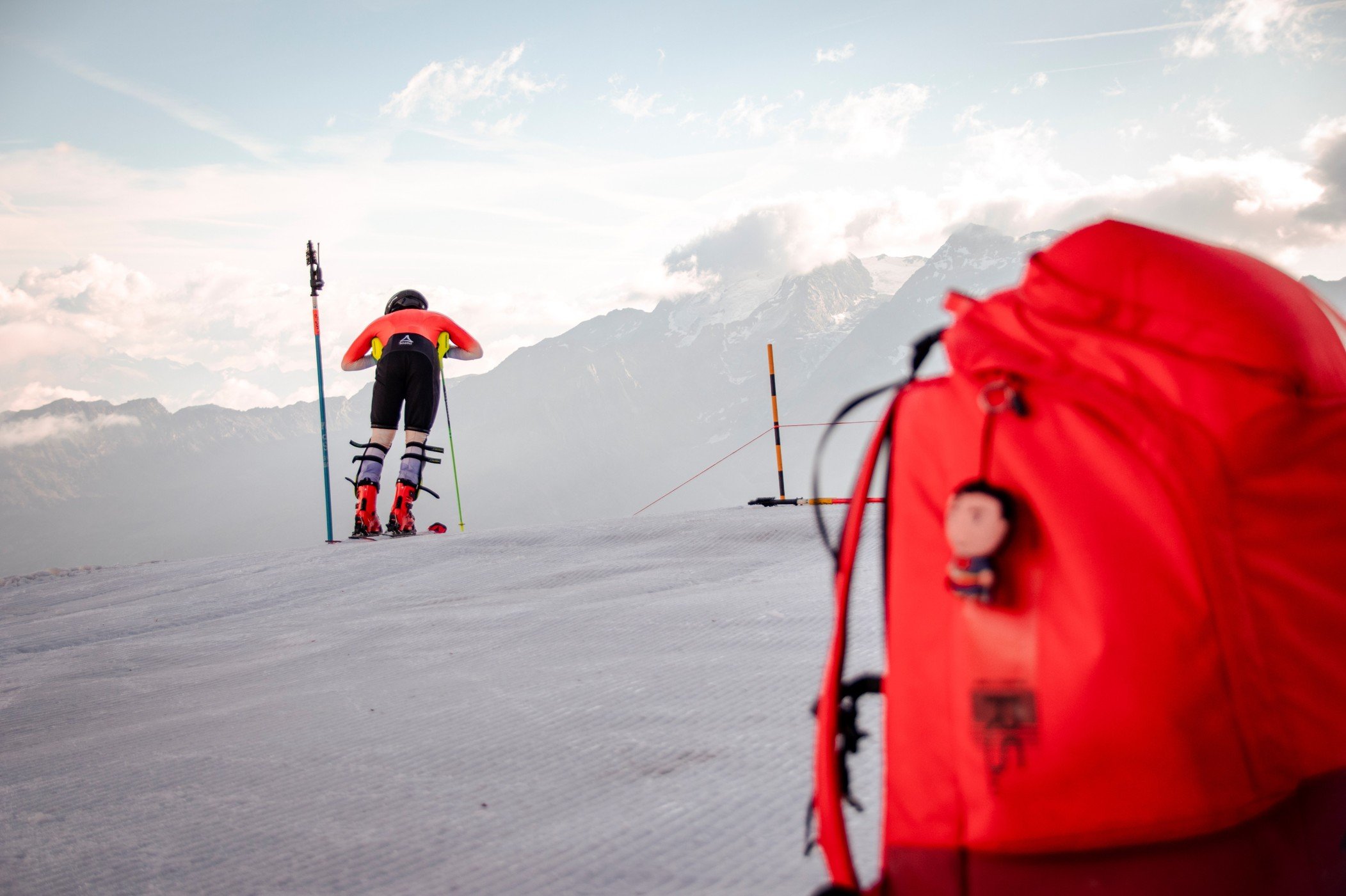 Skifahrer in Rennkleidung steht auf einer Skipiste mit Skistöcken, bereit zum Start. Roter Rucksack im Vordergrund. Berglandschaft im Hintergrund.