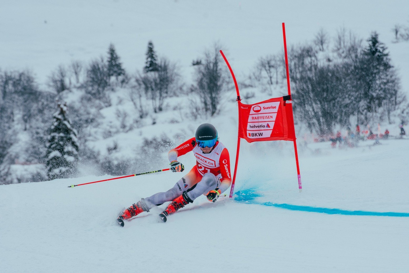 Skifahrer in rotem Rennanzug fährt eine Slalomstrecke mit Torstangen im Schnee, umgeben von verschneiter Landschaft.