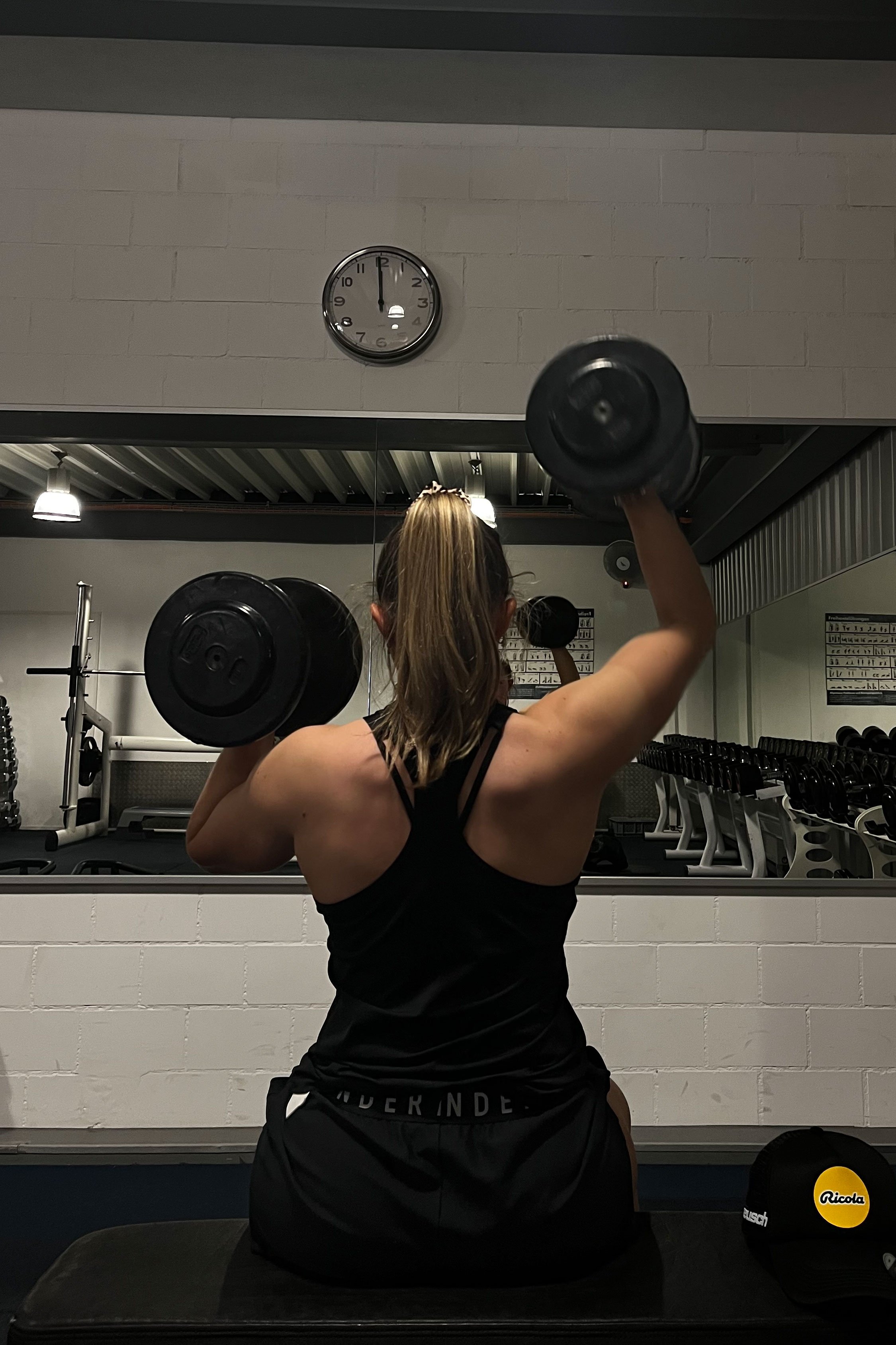Isabella trainiert im Fitnessstudio mit Hanteln, von hinten gesehen, Spiegel an der Wand, Wanduhr zeigt kurz nach 9 Uhr, Sportbekleidung, dunkler Raum, Ricola-Logo auf Mütze im Vordergrund links.