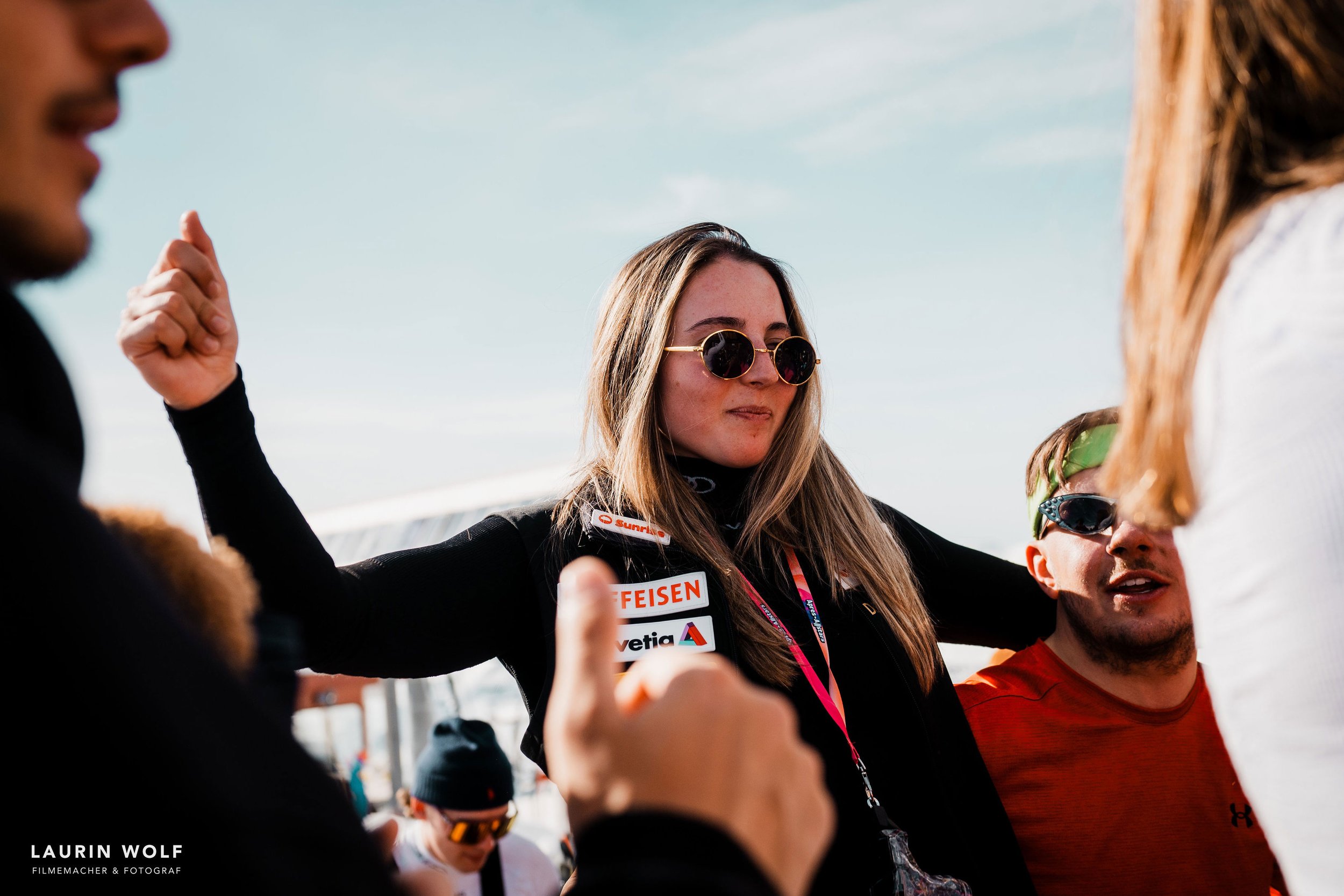 Isabella mit Sonnenbrille und langen Haaren trägt eine schwarze Jacke mit Logos. Sie hebt die Arme in der Luft. Im Hintergrund sind verschwommen andere Personen zu sehen.
