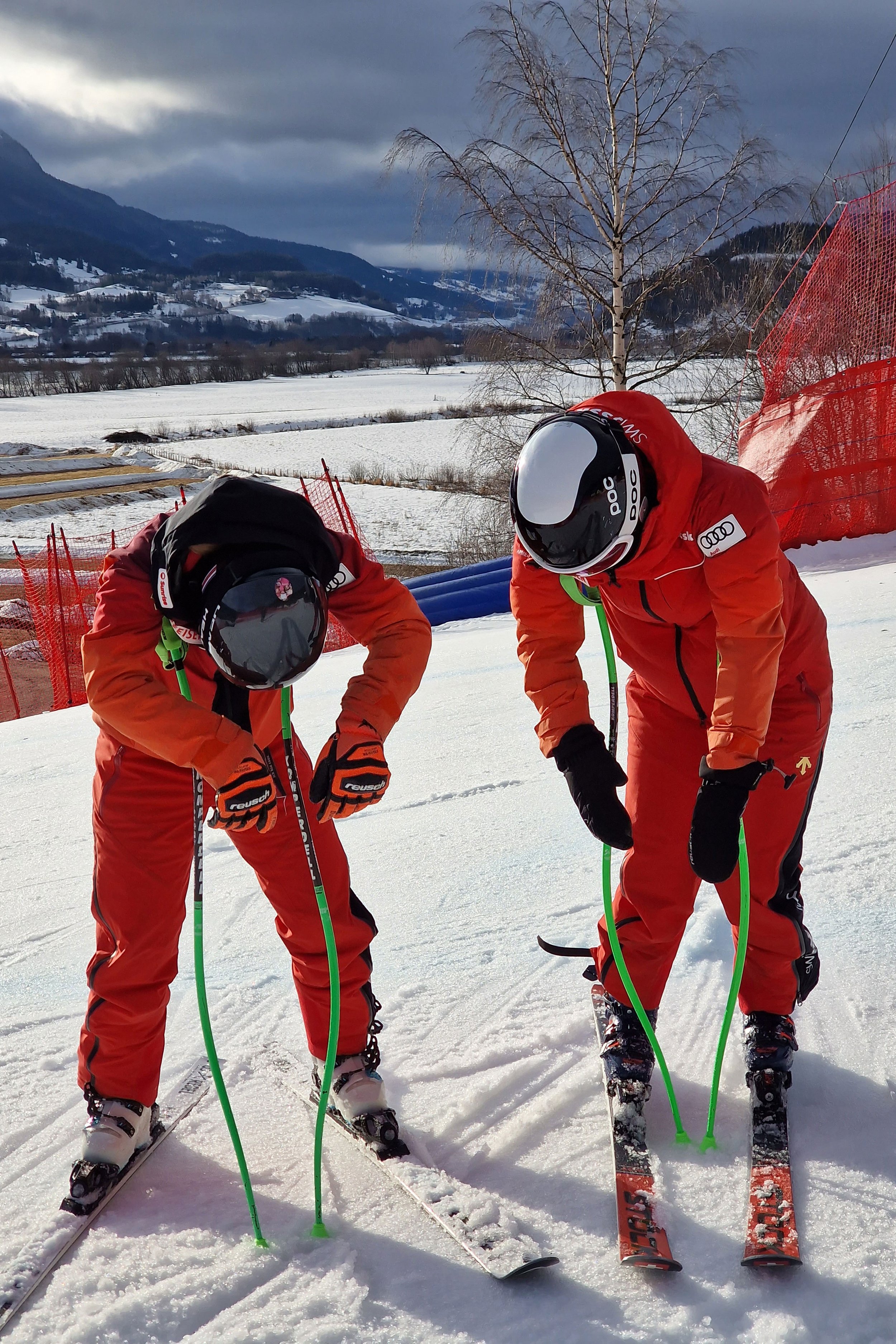 Zwei Skifahrer in roter Skibekleidung beugen sich vor auf einer Schneepiste, halten Skistöcke und tragen Helme. Im Hintergrund ist eine schneebedeckte Landschaft mit Hügeln und Bäumen zu sehen.