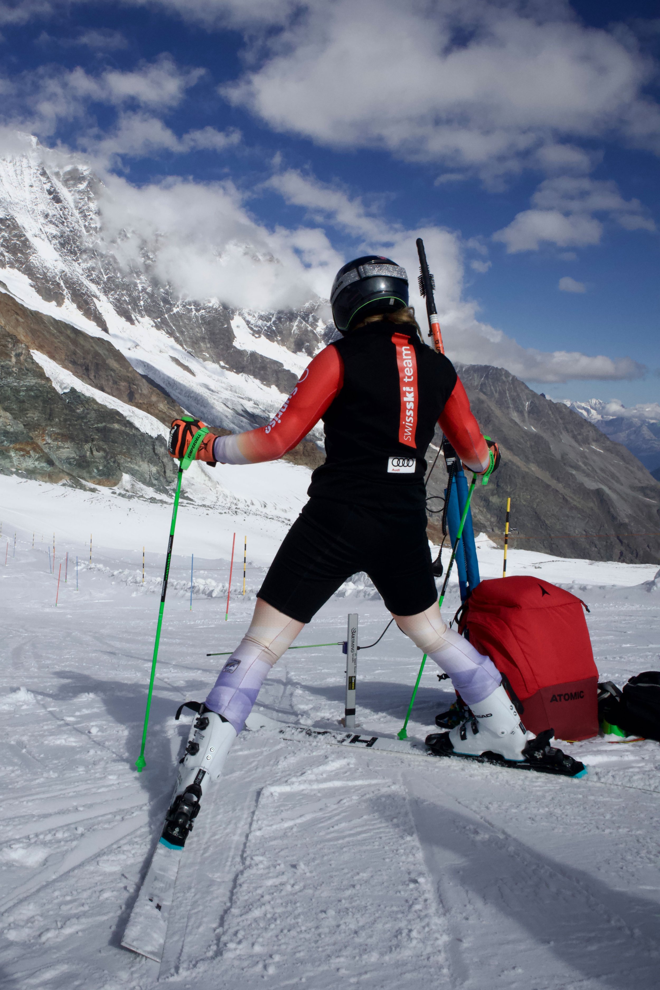 Skifahrerin in roter Jacke mit Skistöcken auf verschneitem Hang vor Bergkulisse.