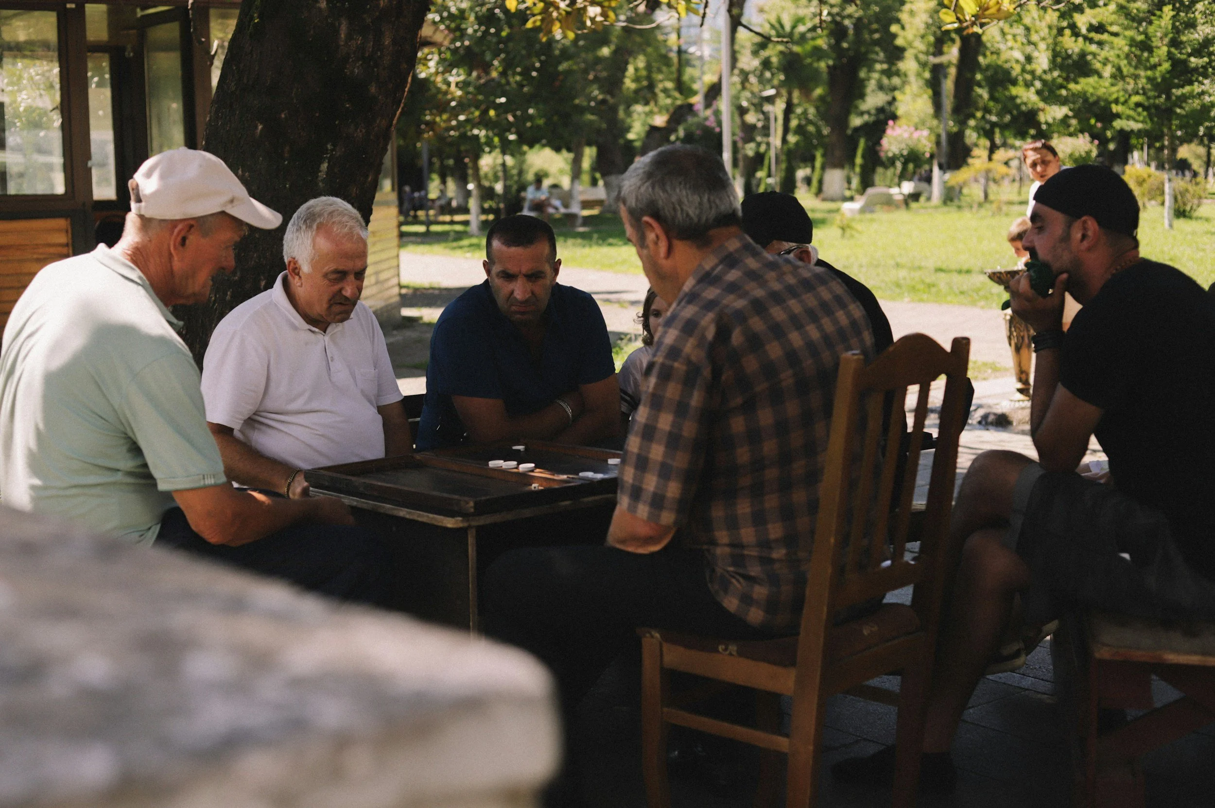 People playing backgammon outdoors under a tree in a park on a sunny day.