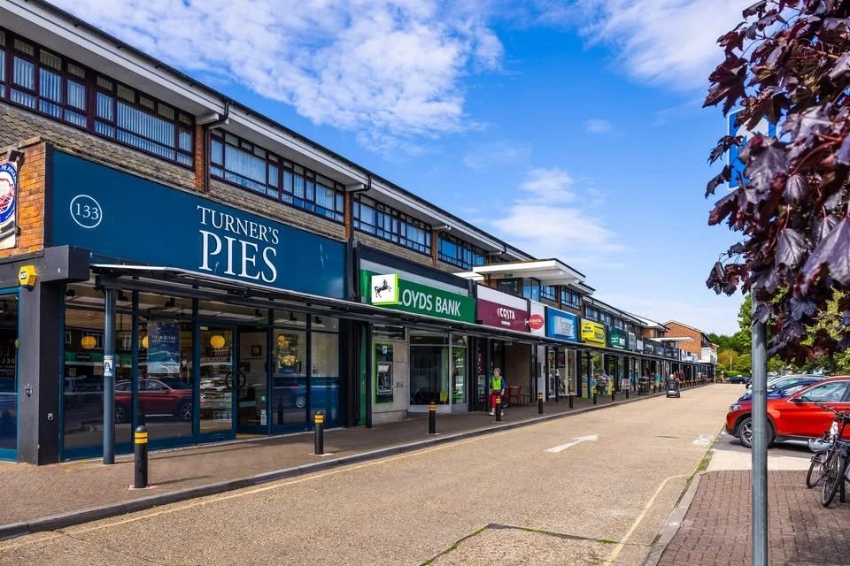 A shopping center with various storefronts, including Turner’s Pies, Lloyds Bank, Costa Coffee, and others, under a clear blue sky with some clouds, parked cars, bicycles, and people walking.