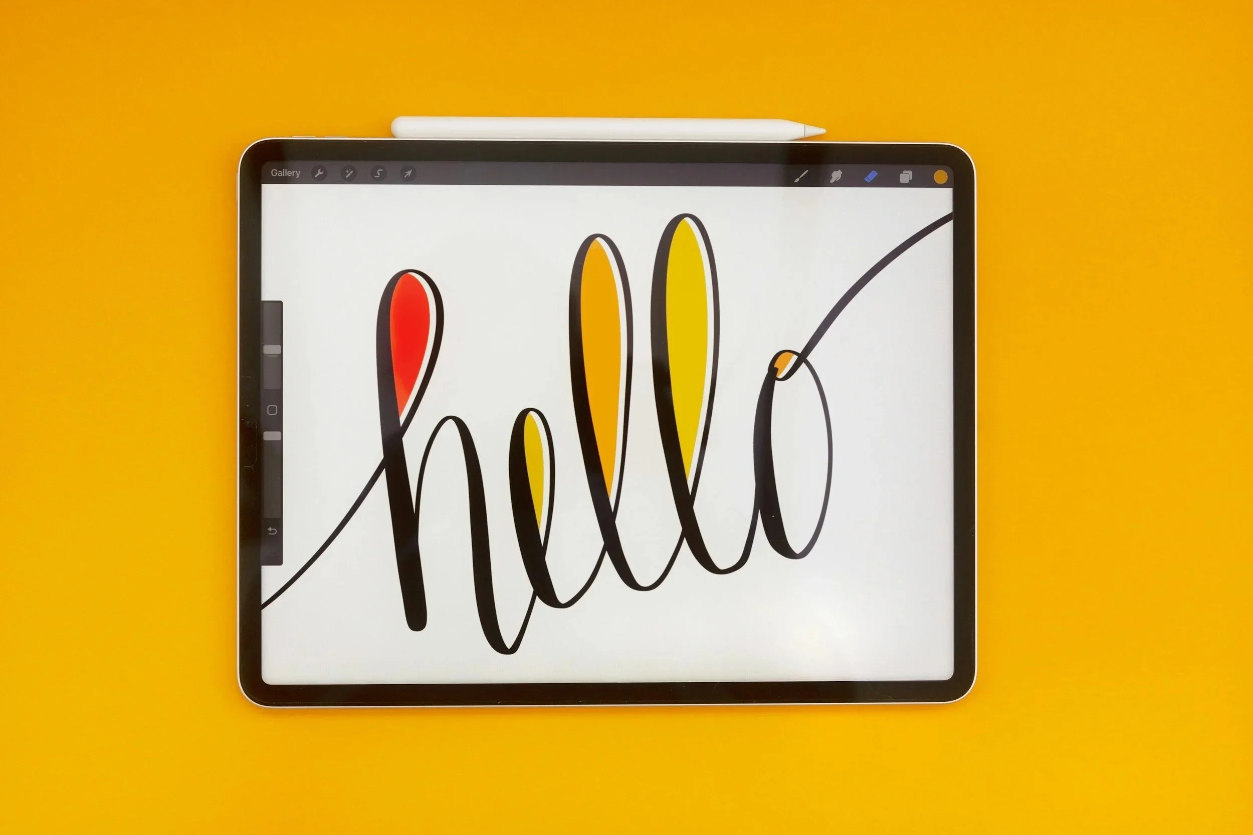 A digital tablet displaying the word "hello" written in black cursive with a yellow, orange, and red background, placed on a yellow surface with a white stylus resting above it.