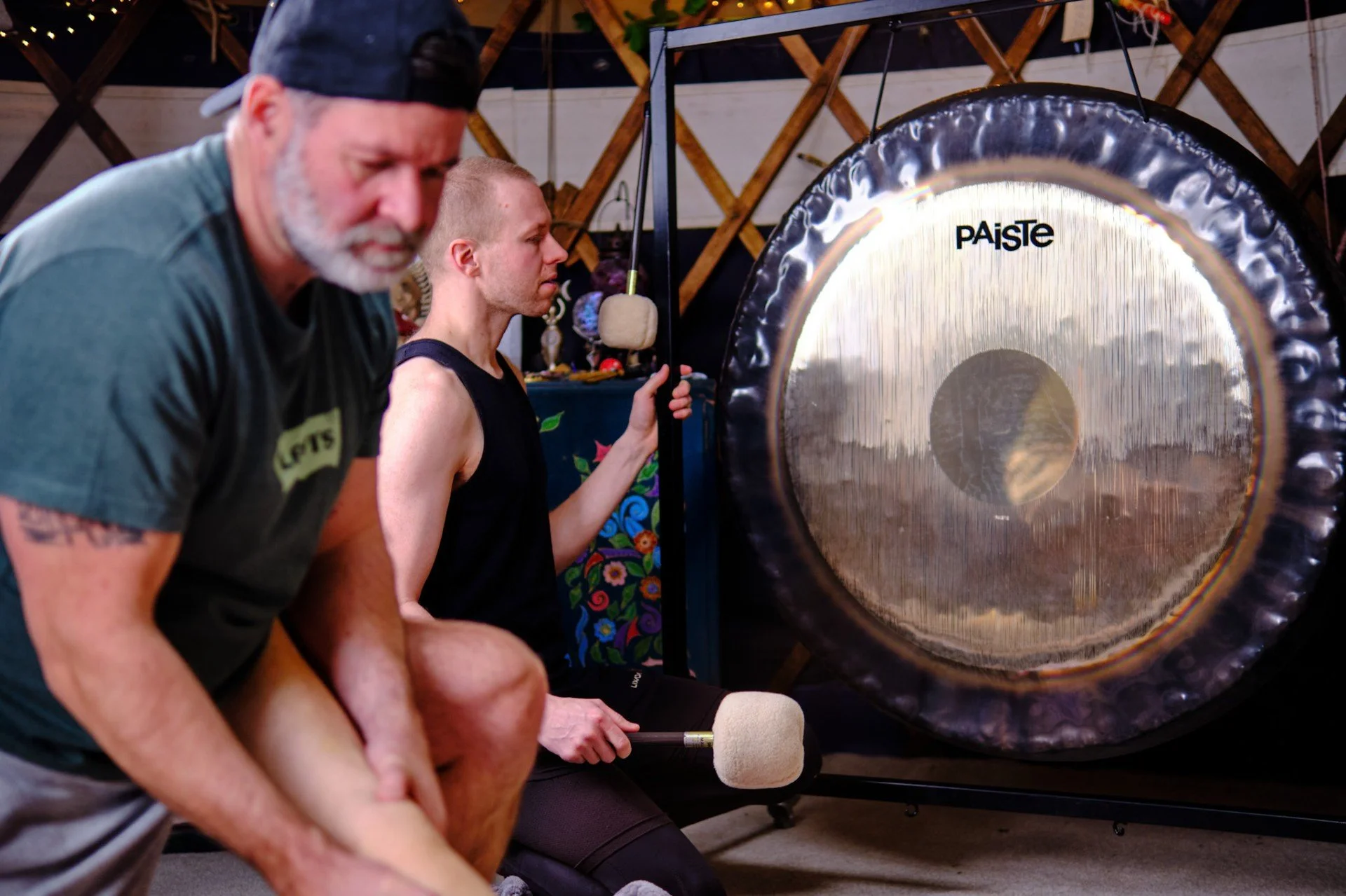 Three men sitting next to a large gong with a wooden frame, with one holding a mallet, in a room with wooden walls and purple decorations in the background.
