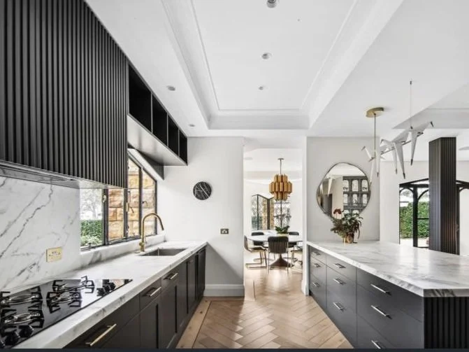 Modern kitchen with black cabinetry, marble backsplash, gold faucet, and a view into a dining area with a wooden chandelier and a round mirror on the wall