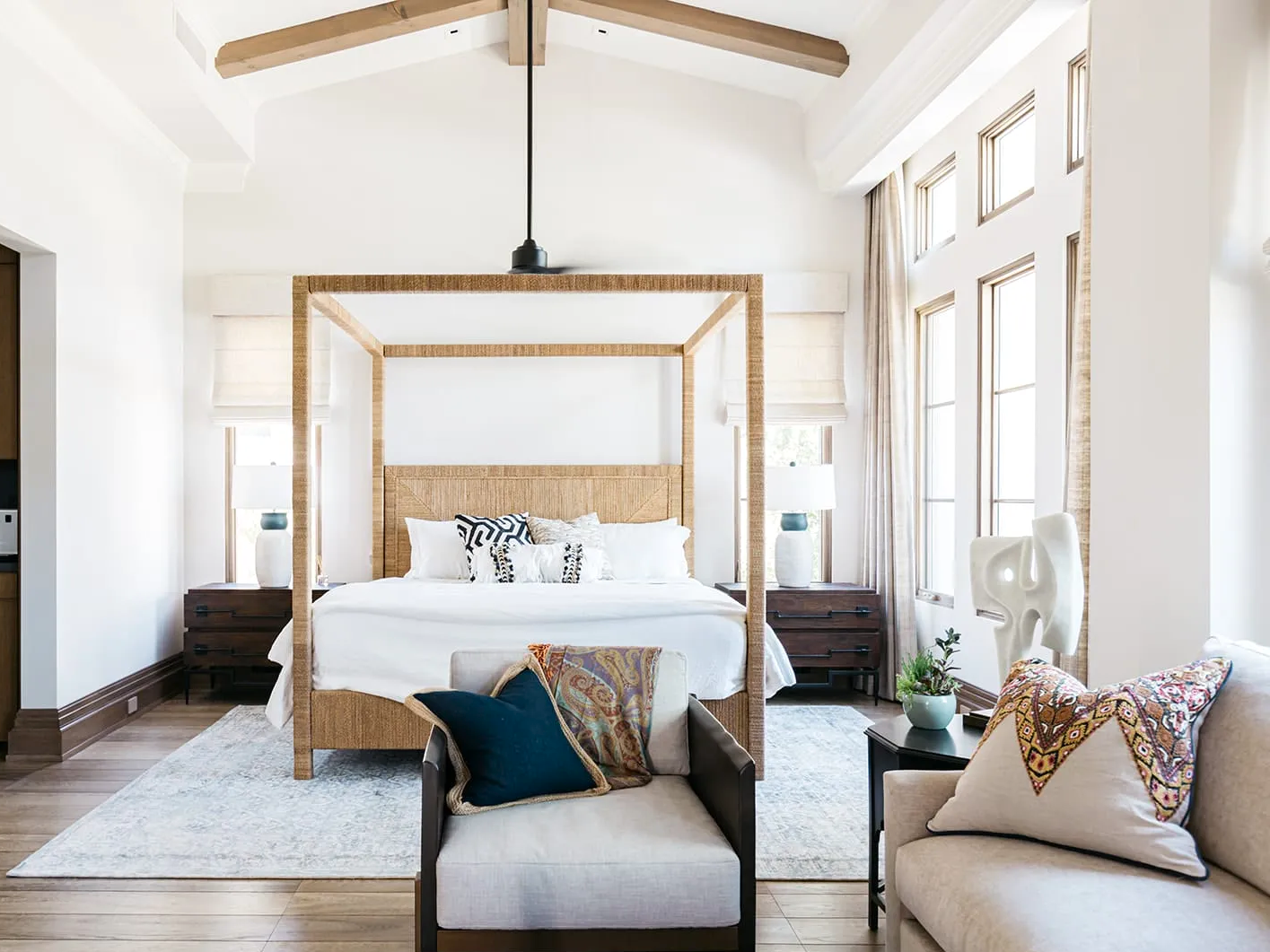 A spacious bedroom with a high vaulted ceiling, large windows, and a king-sized bed with a woven headboard, decorated with patterned throw pillows. There is a four-poster canopy frame over the bed, two nightstands with table lamps, a beige armchair with pillows, and abstract art sculpture beside the window. Light, neutral colors and natural light create an open, airy atmosphere.