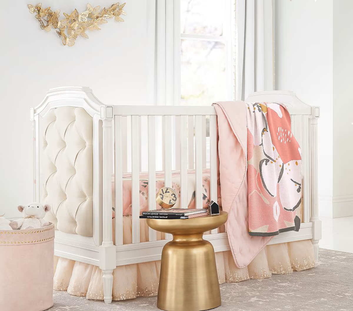 A white crib with tufted headboard and pink bedding, decorated with pink and floral blankets hanging over the side, in a bright nursery with a window, a gold bedside table with books and a clock, and a butterfly wall decoration.