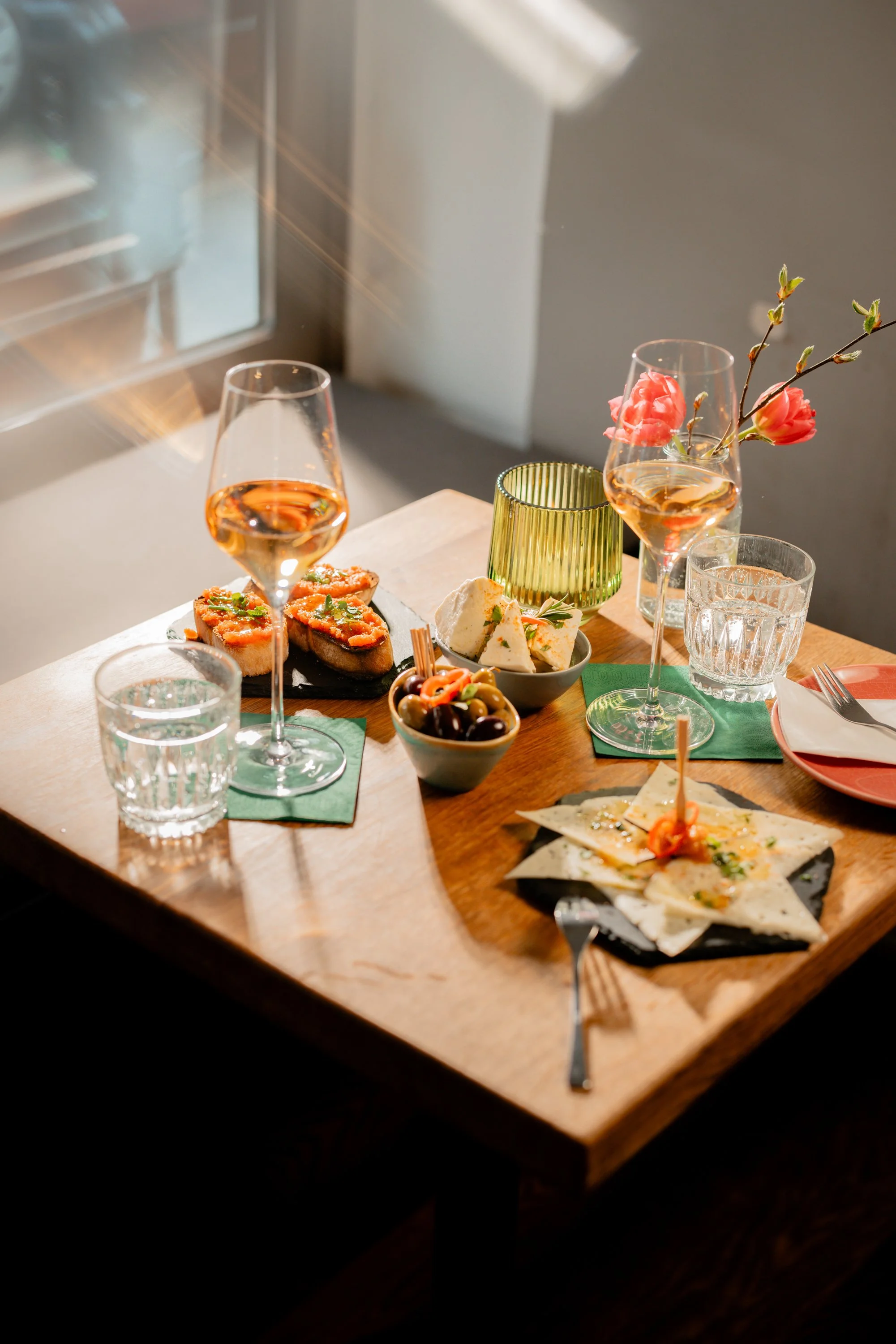 Ein gedeckter Tisch mit zwei Gläsern Weißwein, Tellern mit Pan con Tomate, Manchego Käse, eingelegte Oliven und Blumen in einer Vase. Die Sonne strahlt durch das Fenster auf den Tisch.