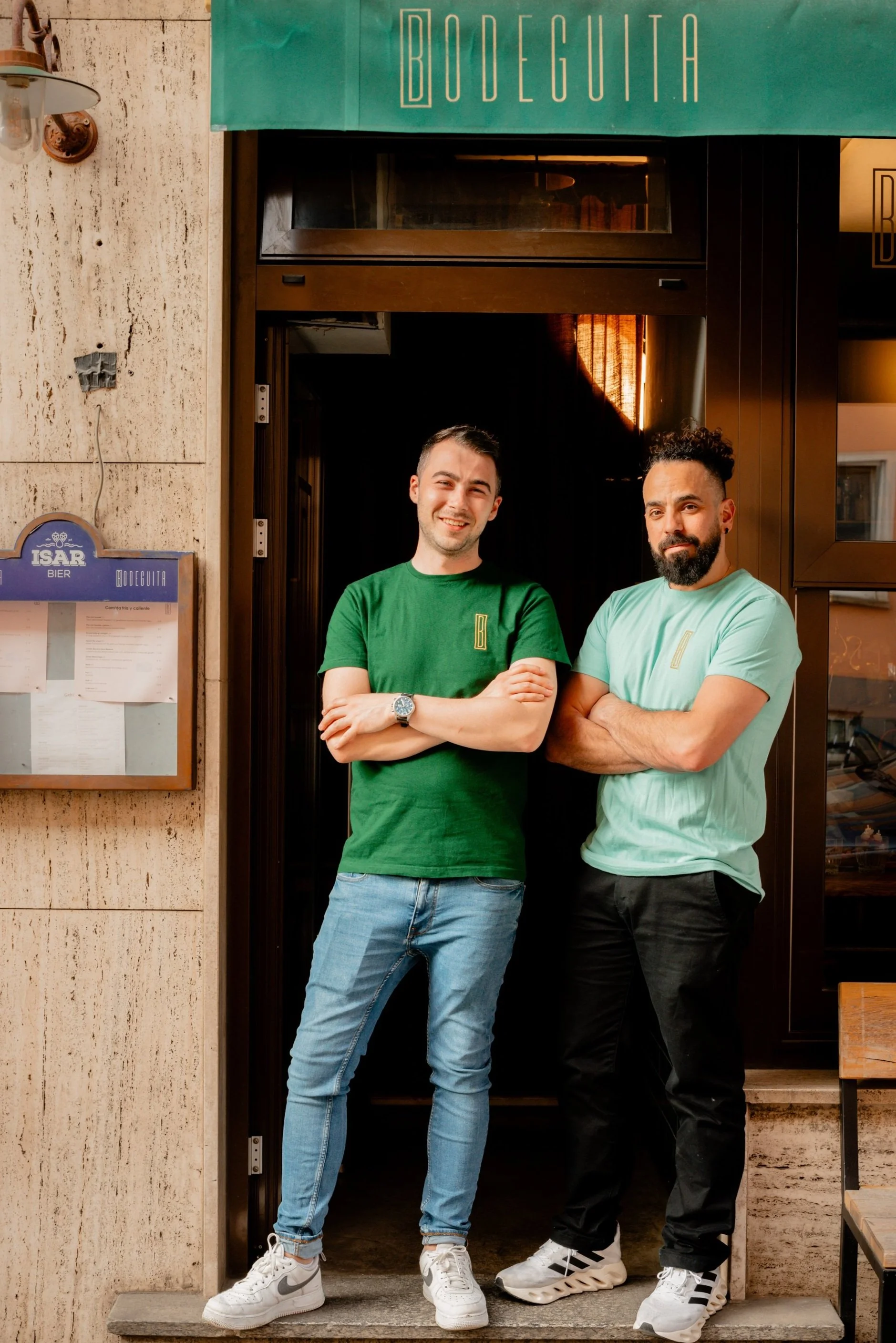 Zwei Männer stehen vor einem Restaurant mit dem Schild "Bodeguita" über dem Eingang. Der linke Mann trägt ein grünes T-Shirt und Jeans, der rechte Mann ein hellgrünes T-Shirt und schwarze Hosen. Beide lächeln und stehen mit verschränkten Armen.