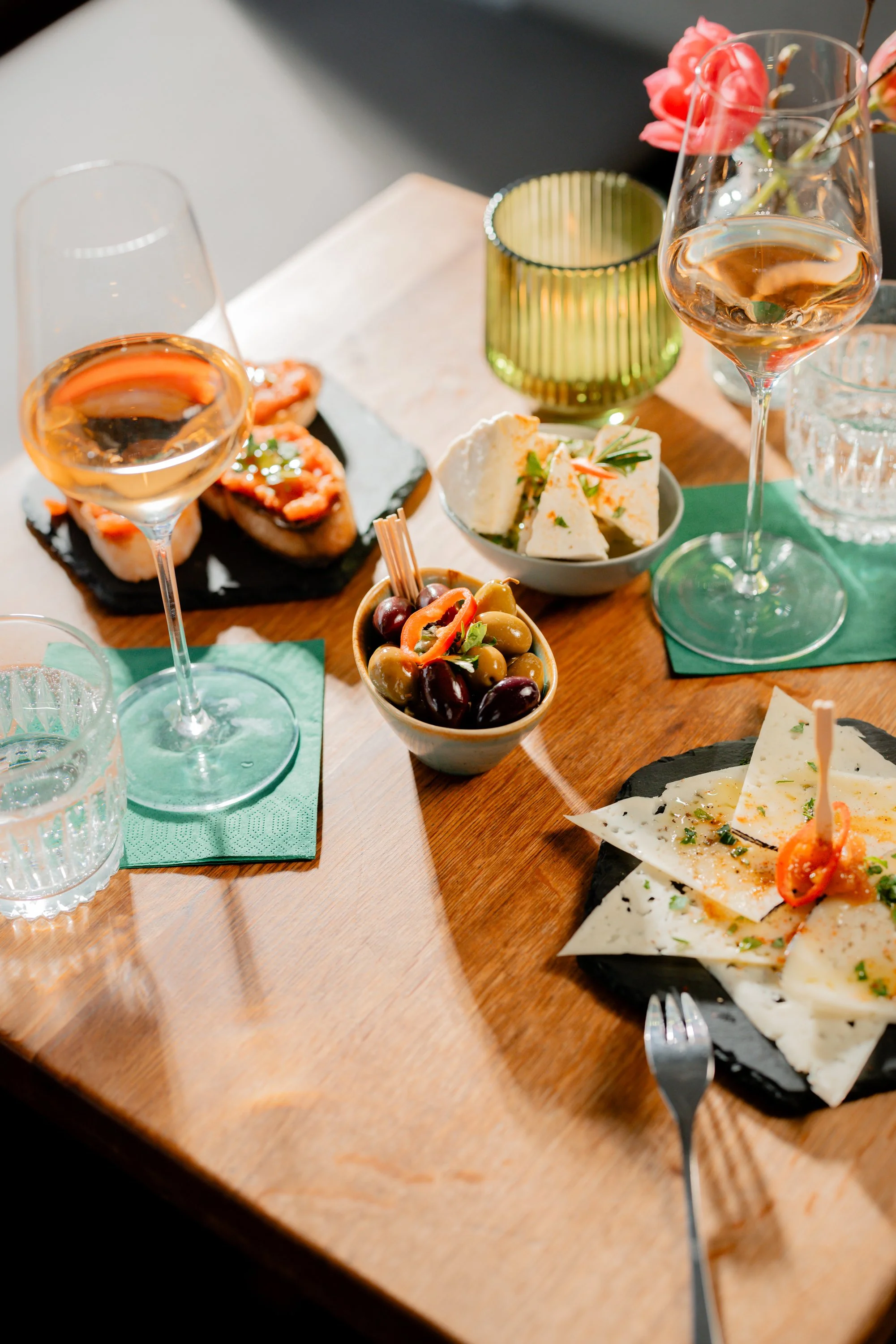 Ein Tisch mit Tapas, darunter Oliven, Manchego Käse, Pan con Tomate und zwei Gläser Wein. Eine grüne Kerze und ein Wasserglas sind ebenfalls zu sehen.