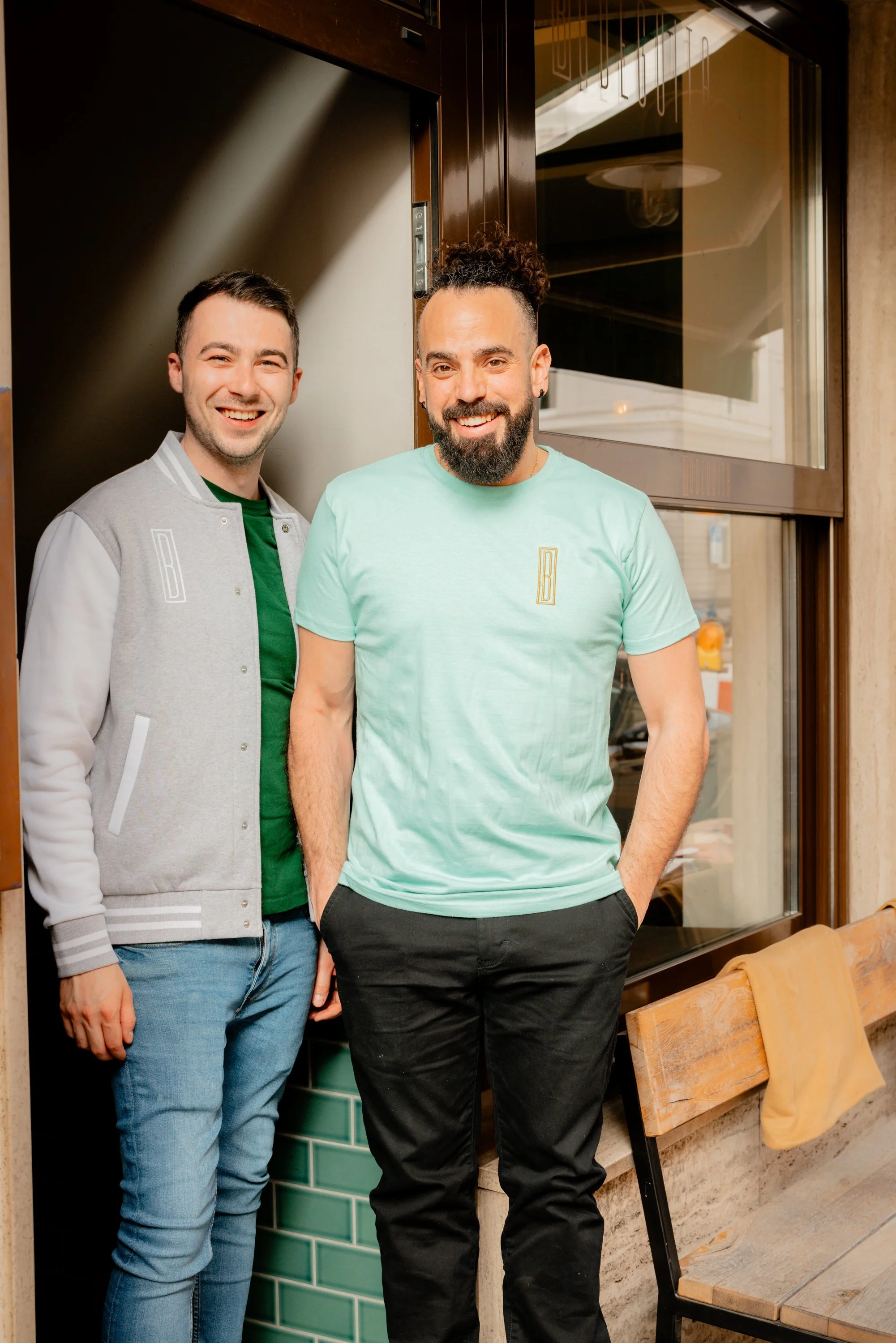 Zwei lächelnde Männer stehen in einem Türrahmen. Einer trägt eine graue College-Jacke , ein grünes T-Shirt und Jeans, der andere ein mintgrünes T-Shirt und schwarze Hose.