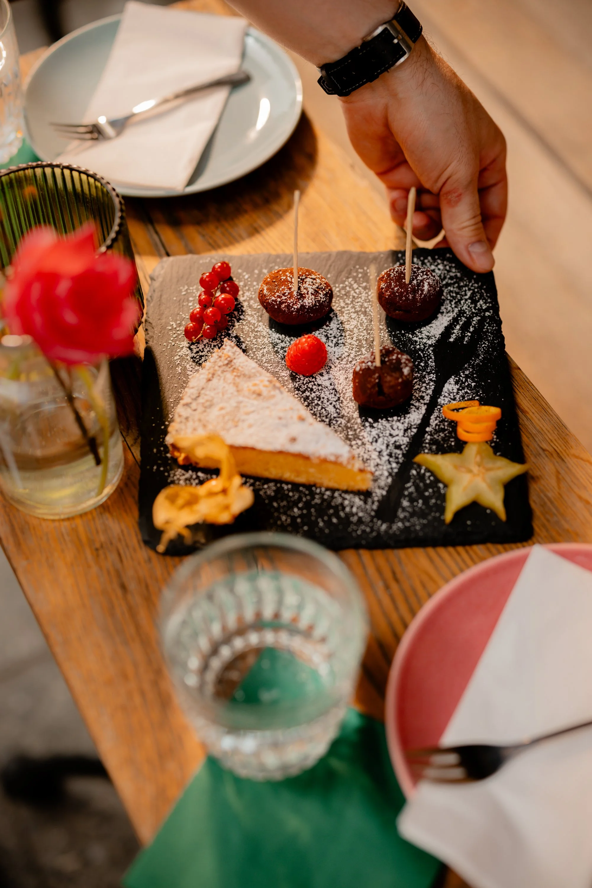 Schwarzplatte mit verschiedenen Desserts, darunter ein Stück Kuchen mit Puderzucker, kleine Schokoladenkugeln auf Spießen, Johannisbeeren, Himbeere, getrocknete Frucht, Sternfrucht und Orangenspirale. Eine Hand hält die Platte. Im Hintergrund sind ein Glas, ein Teller mit Besteck und eine Vase mit Blume zu sehen.