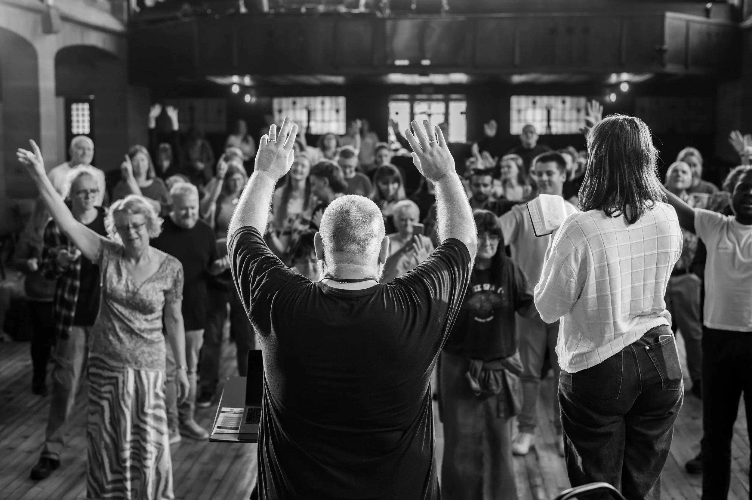 A group of people standing in front a man whose back we see, that appears to have his hands lifted in blessing towards the large group in front of him.